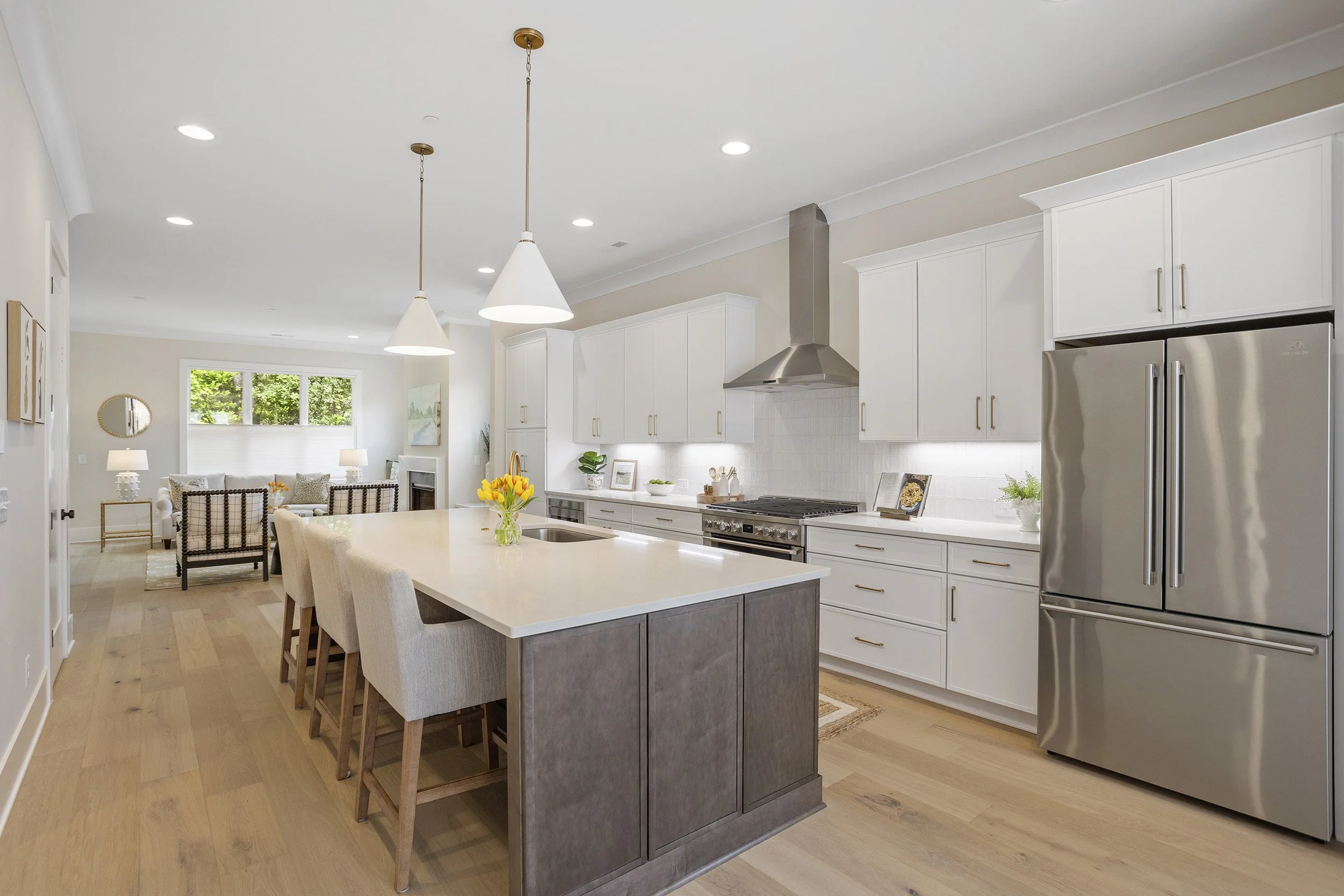 Open-concept kitchen with white cabinets, stainless steel refrigerator and stove, central island with darker wood base, pendant and recessed lighting, and a view into the living room with a beige couch and round mirror on the wall.