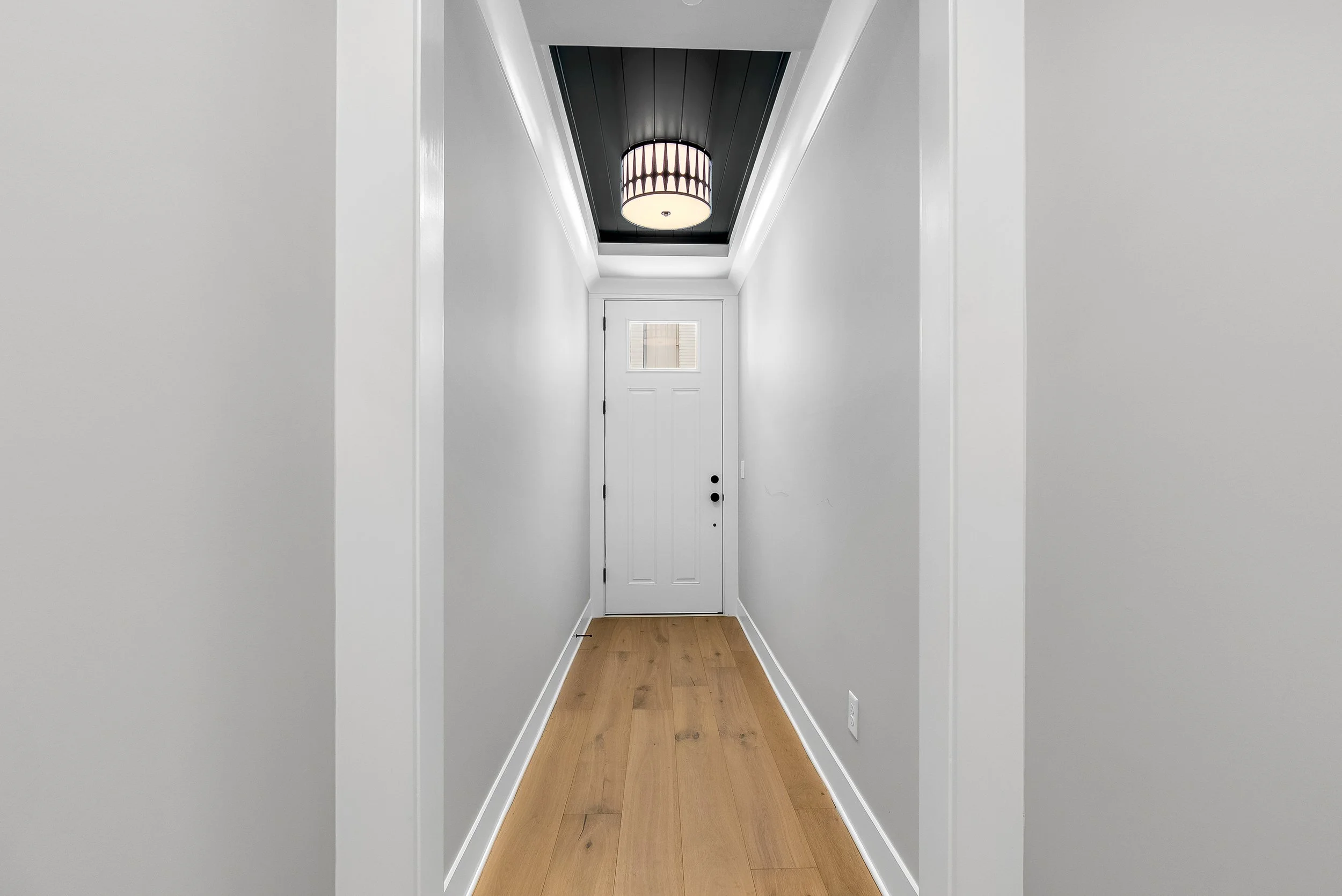 A narrow hallway with white walls, a wooden floor, and a white door at the end. There is a window on the door, a ceiling light fixture with a cylindrical shade, and white crown molding along the ceiling edges.