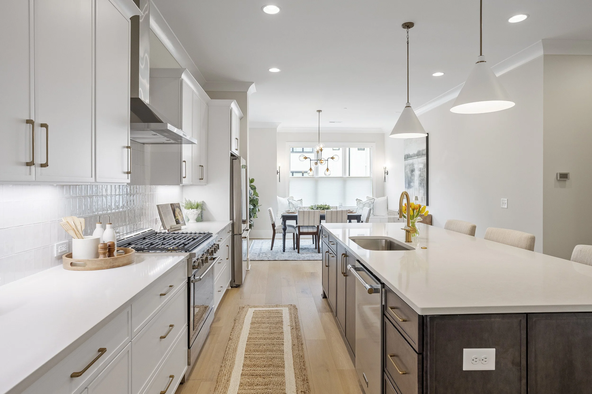 A modern, bright kitchen with white cabinets, a large island, pendant lights, and a view of a dining and living area with large windows.