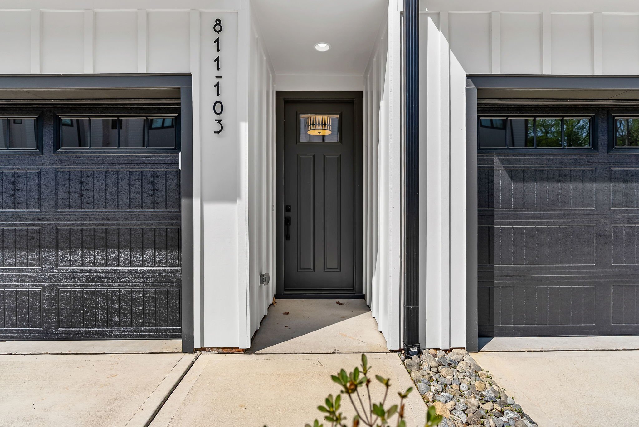 View of a modern house entrance with dark front door, white trim, and black house number 8311. The entrance is framed by a covered porch, with garage doors on either side, and small rocks along the walkway.