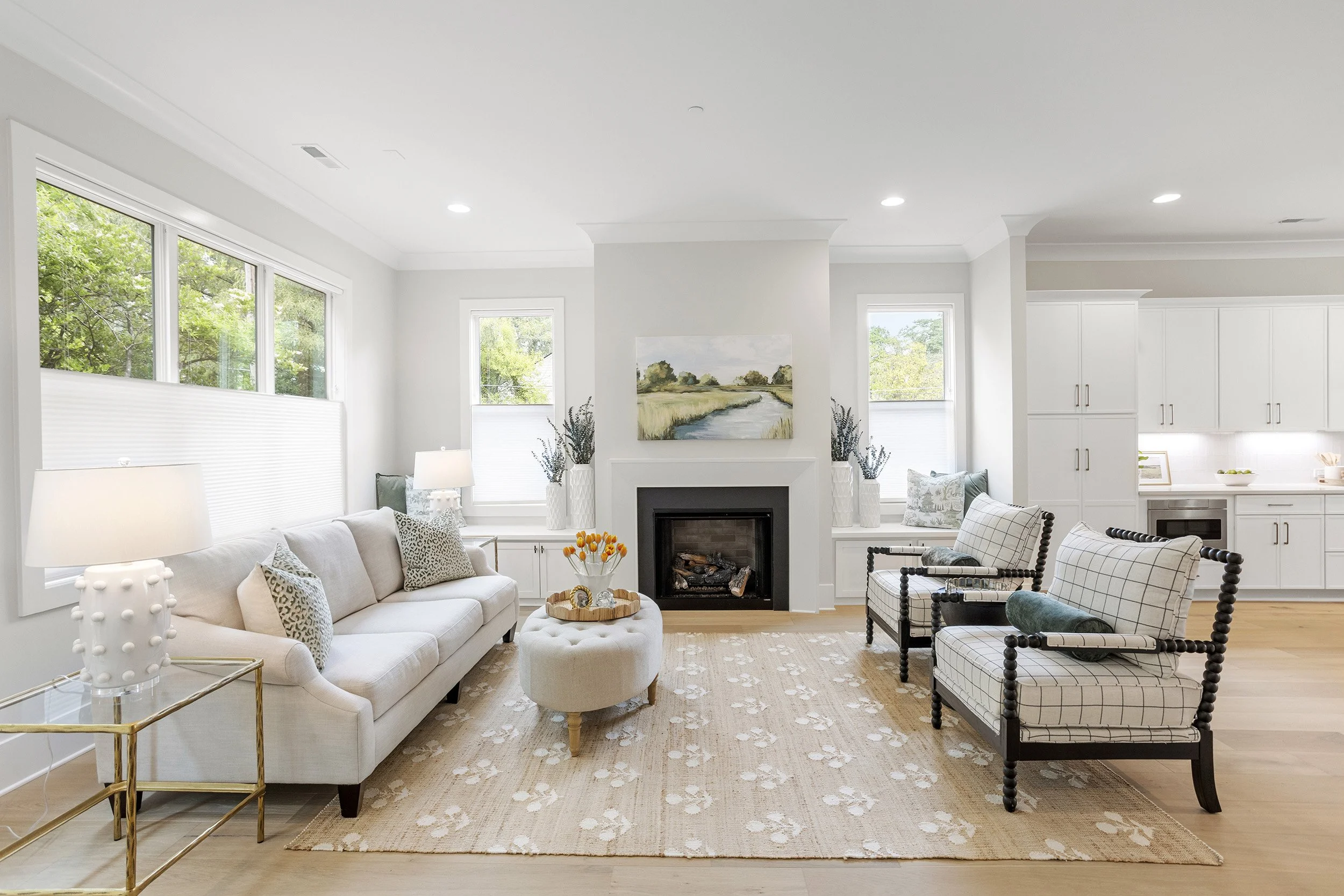 Bright living room with white walls, large windows, a beige sofa with cushions, a fireplace with artwork above, patterned armchairs, a beige rug, and decorative items including lamps and vases with flowers.