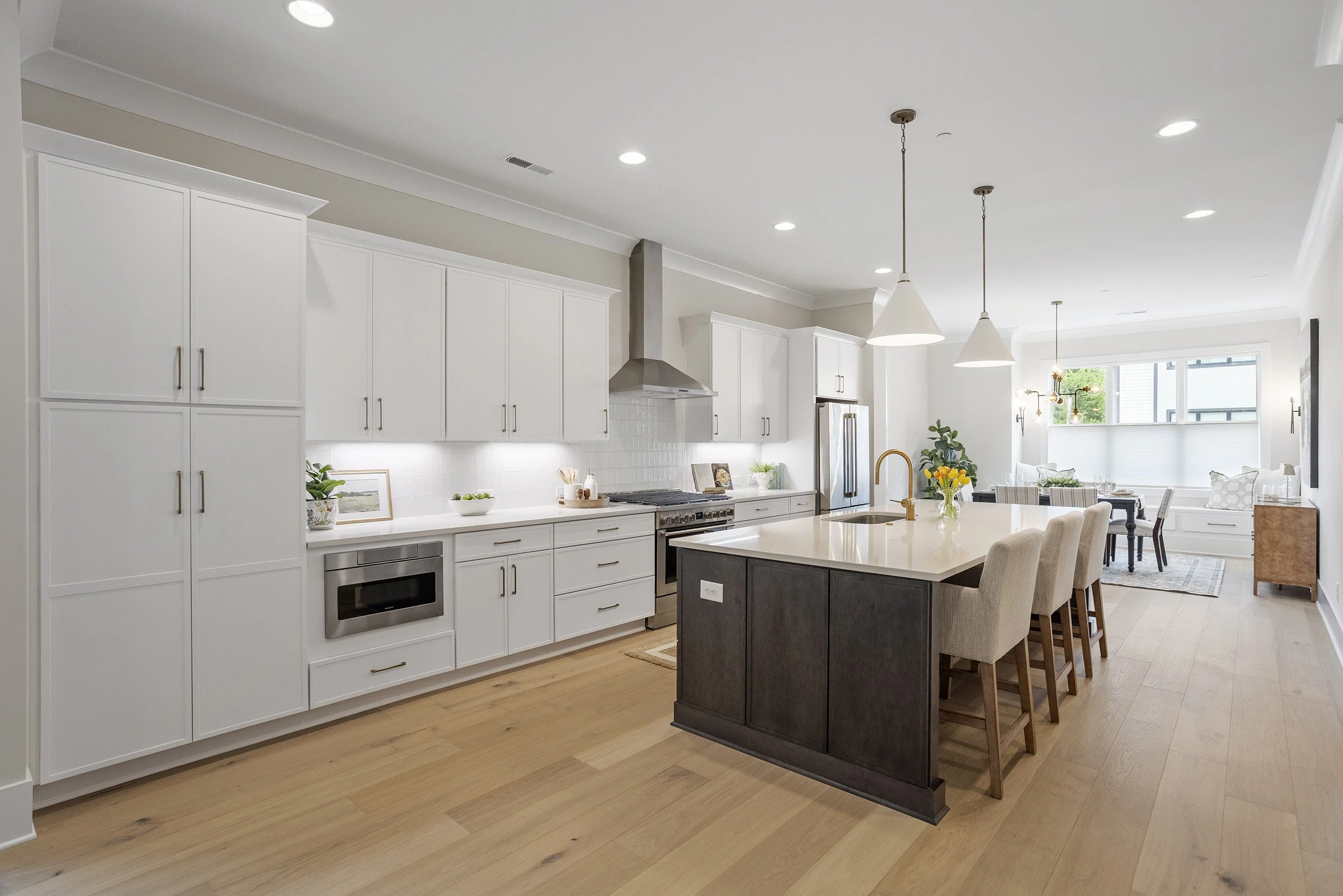 Modern, open-concept kitchen and dining area with white cabinets, stainless steel appliances, a kitchen island with a dark base, pendant lights, and a dining table with chairs. Large windows and hardwood flooring enhance the space.