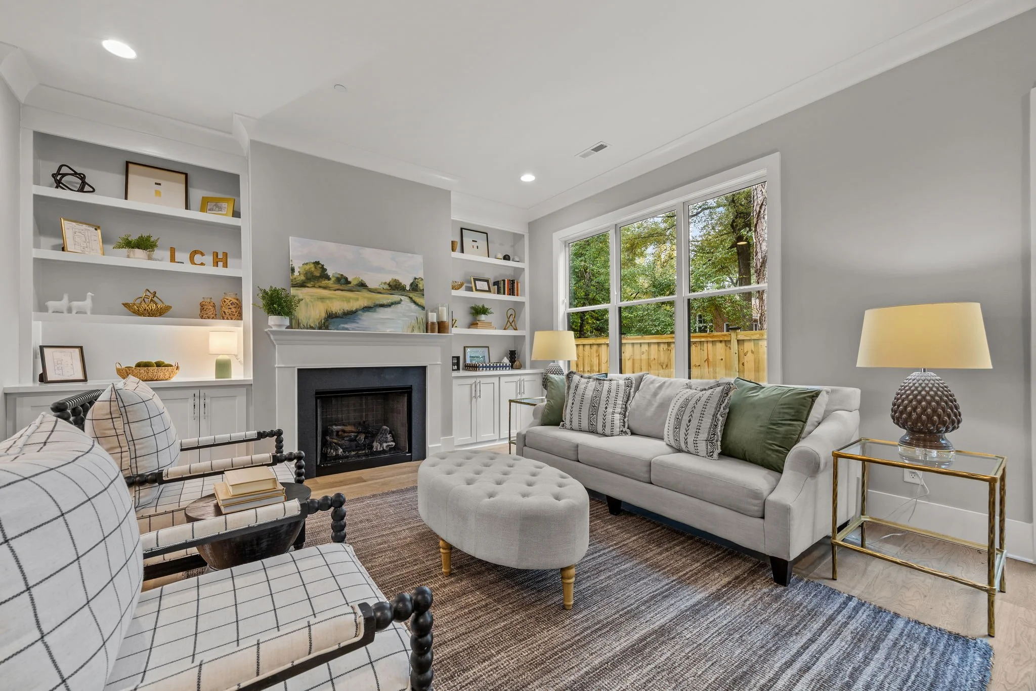Living room with gray walls, white built-in shelves, a fireplace, large window showing outdoor trees, white sofa with patterned pillows, plaid armchairs, table lamps, and a textured round side table.