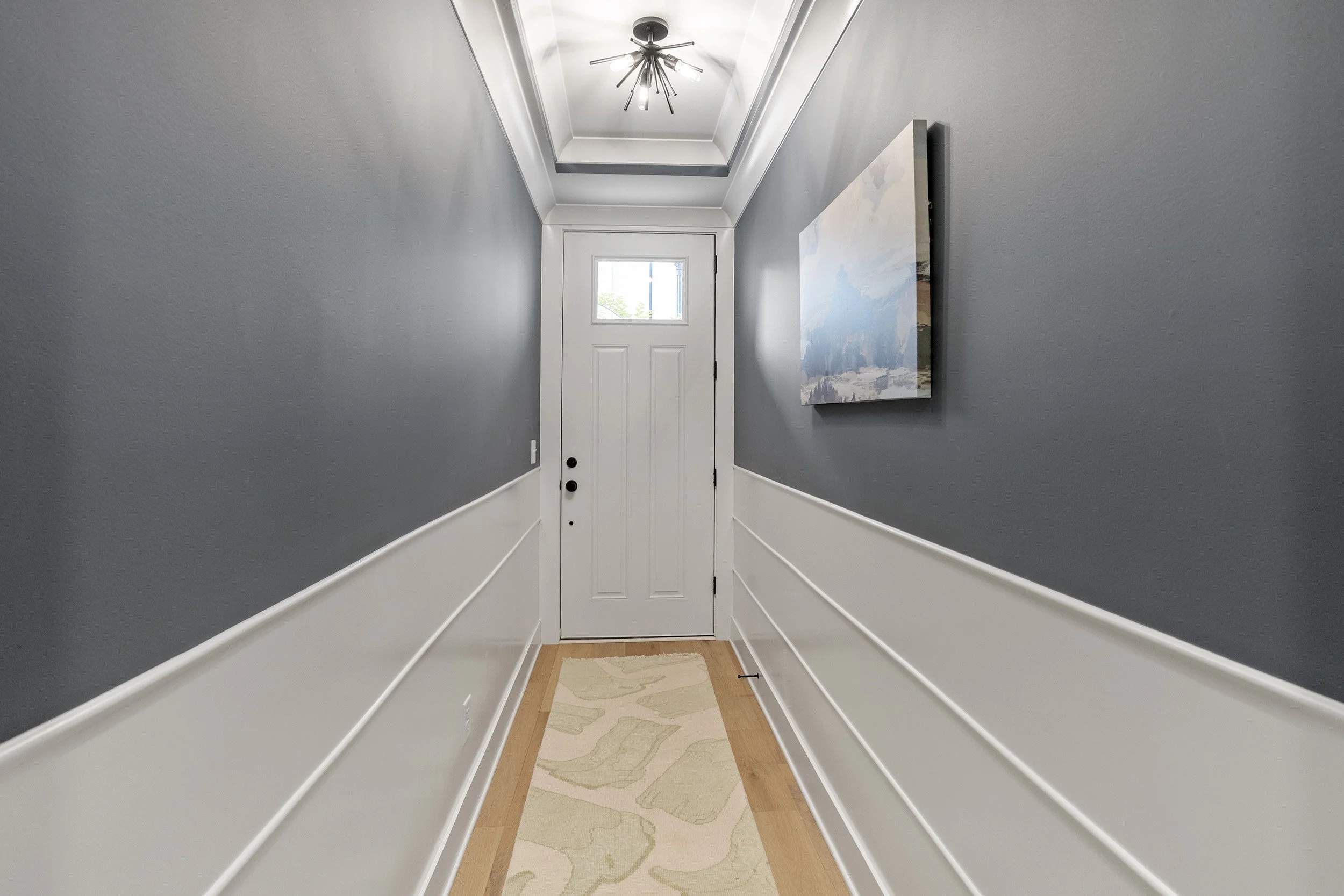 A hallway with gray walls, white trim and molding, a front door with a window, a hanging light fixture, a painting on the right wall, a beige rug with a floral pattern, and a hardwood floor