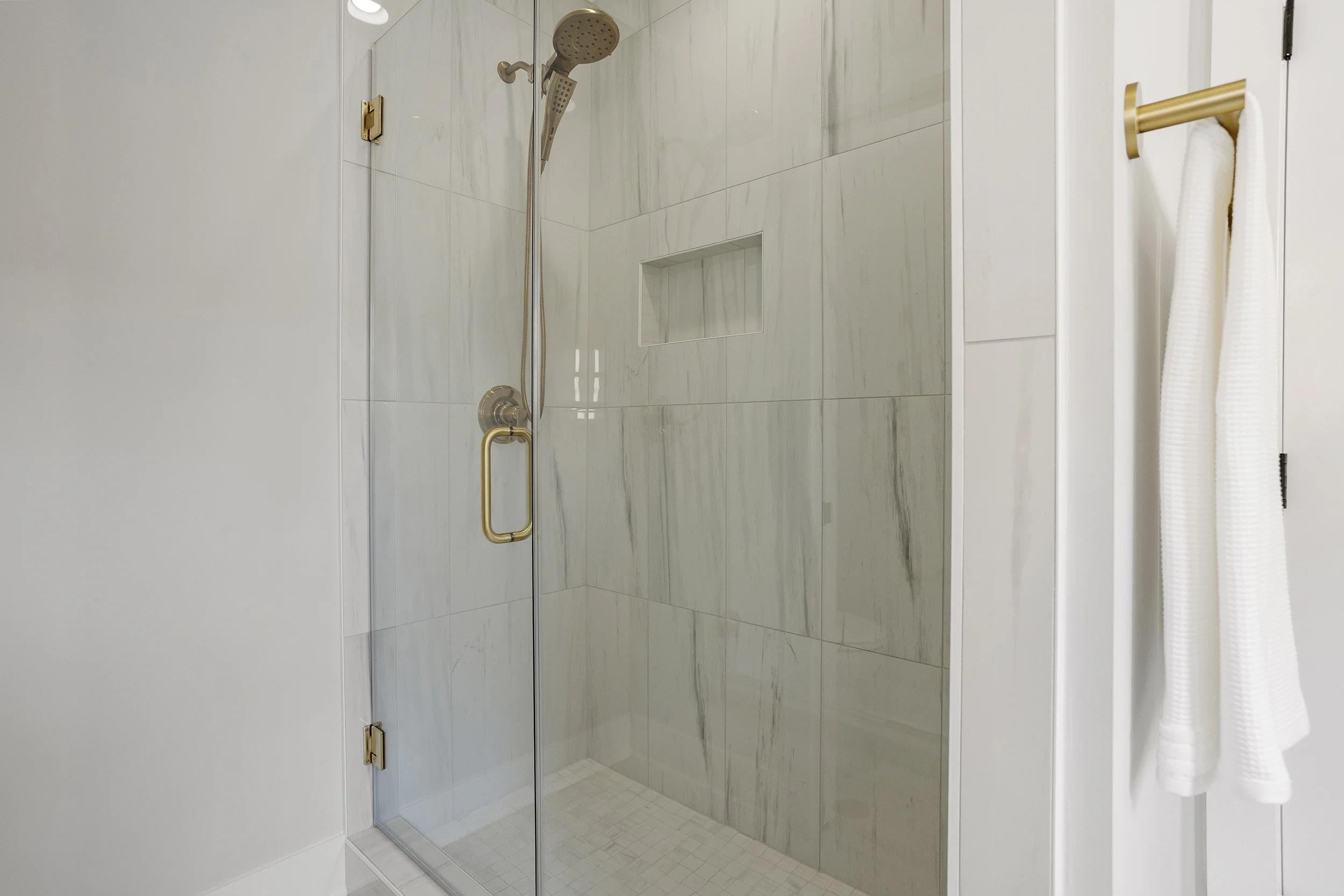 A modern shower with glass doors, a gold handle, a wall-mounted showerhead, and a built-in niche in the tiled wall.