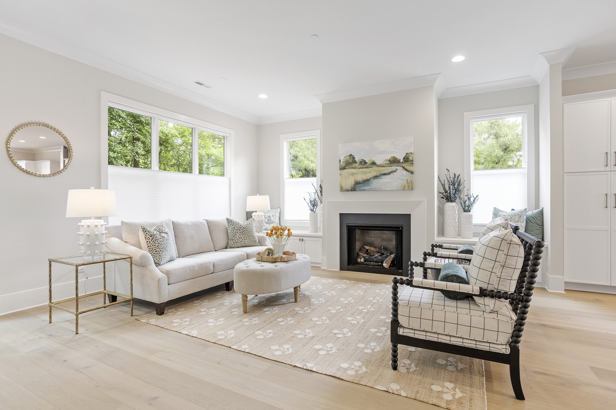 Bright living room with large windows, white walls, a beige sofa with cushions, a fireplace, a round ottoman with a tray, a glass side table, a plaid armchair, and decorative vases and paintings.