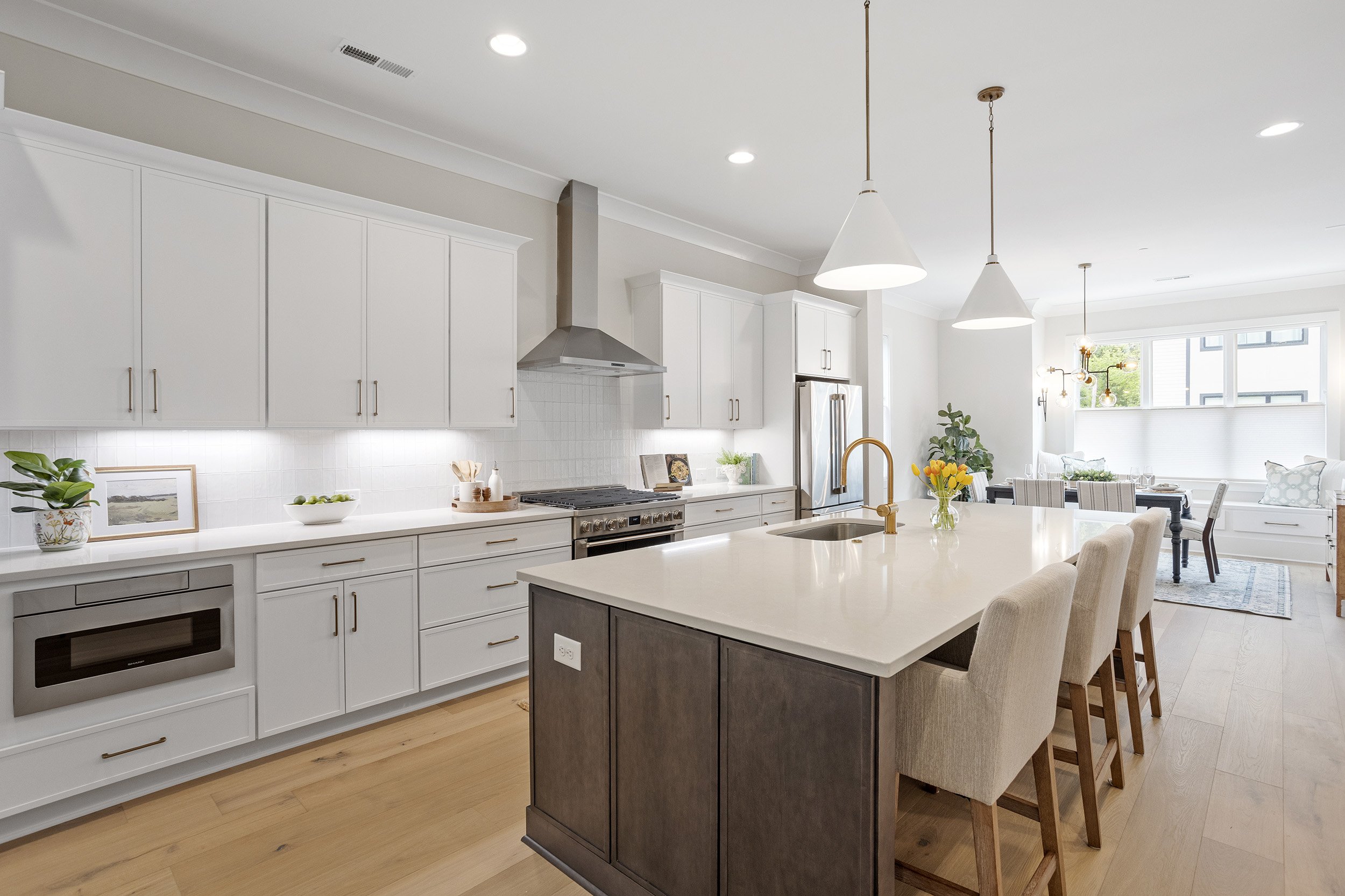 Modern open-concept kitchen with white cabinets, stainless steel appliances, a large island with a dark base, and bar stools. The space is bright with natural light, featuring pendant lights and a dining area with a window view.
