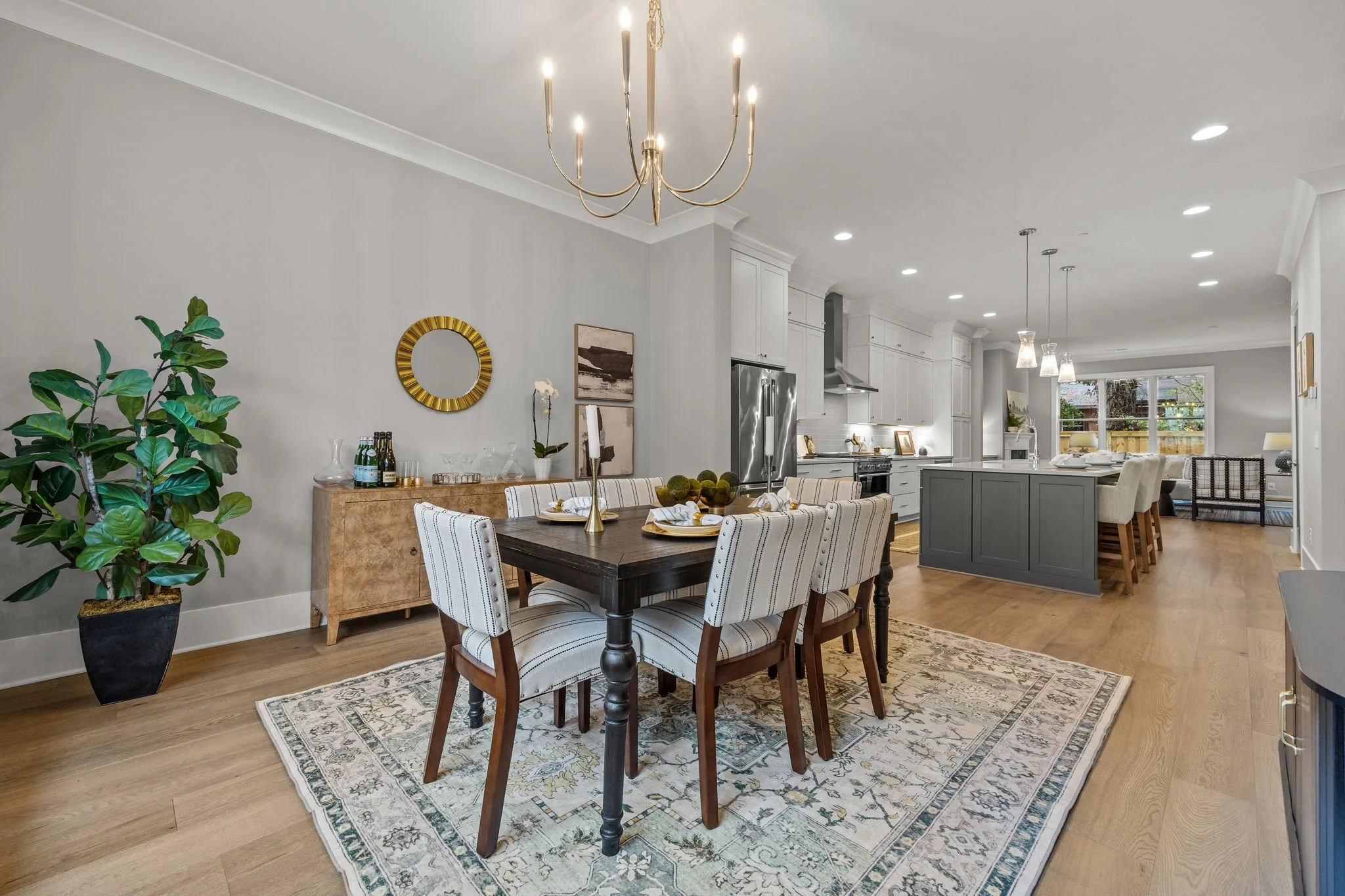 A spacious, modern dining area with a dark wooden table, striped upholstered chairs, a patterned rug, a large green potted plant, and a sideboard with bottles and glasses. The open kitchen in the background features white cabinets, stainless steel ap