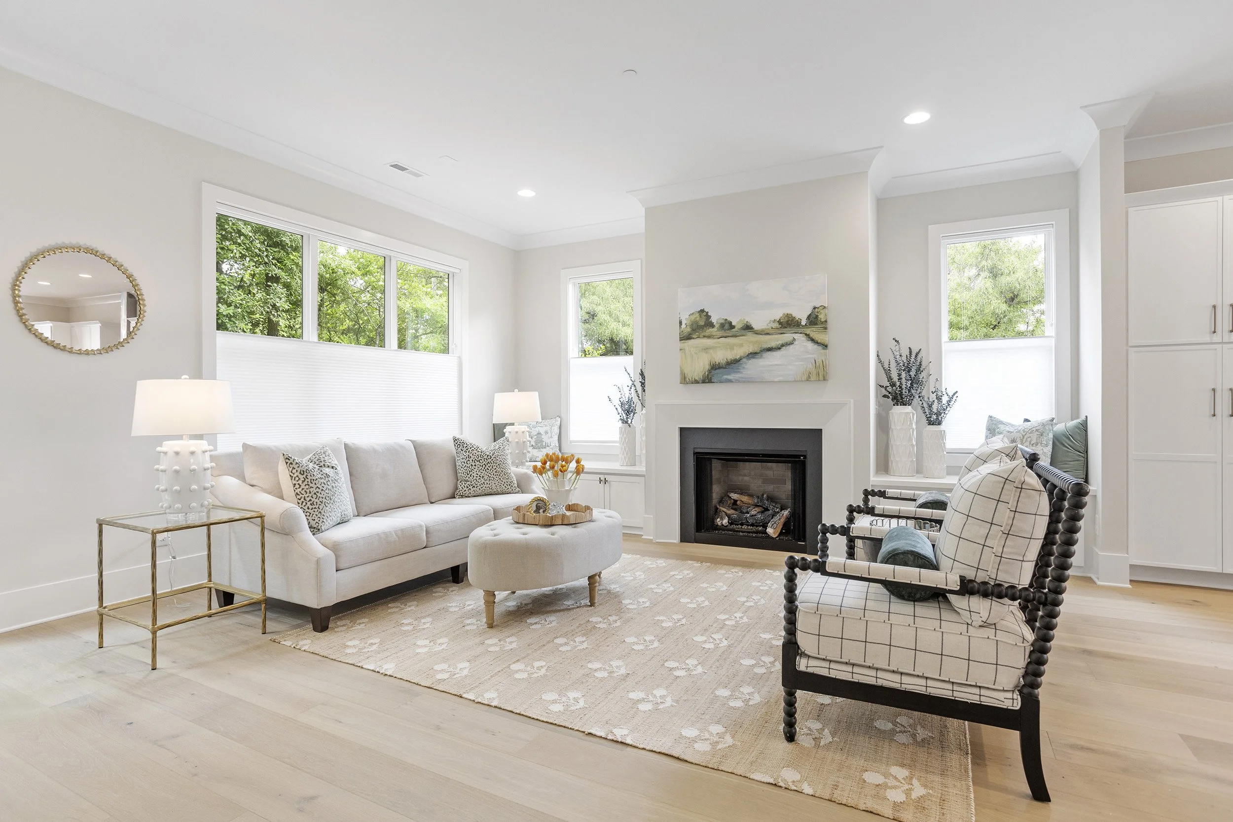 Bright living room with white walls, large windows, white sofa with patterned pillows, black and white patterned armchair, coffee table, fireplace, and decor including two lamps, plants, and artwork.