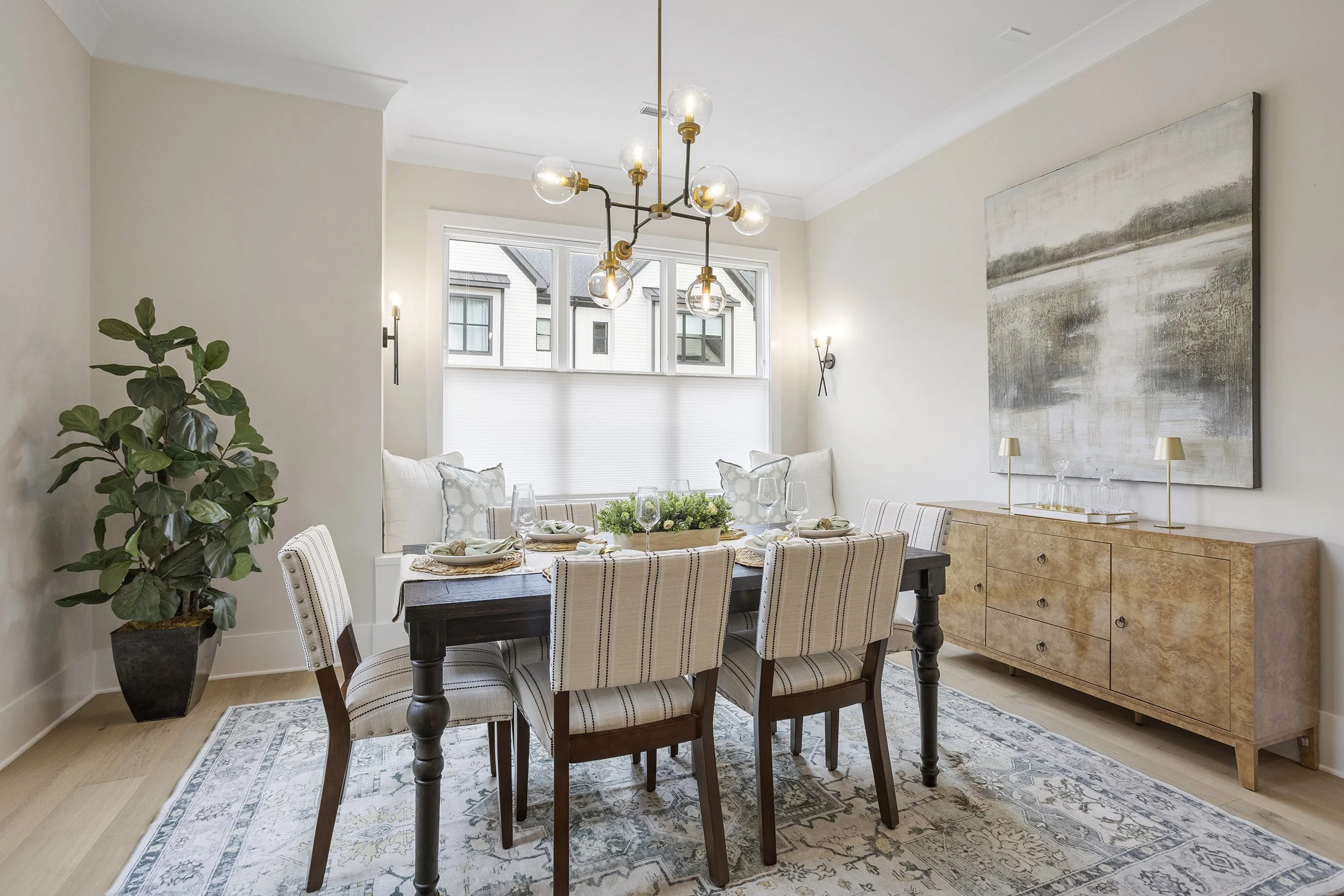 A dining room with a dark wood table, six upholstered chairs with striped fabric, a large abstract painting on the wall, a wooden sideboard with lamps, a tall potted plant, windows with blinds, a chandelier, and part of a rug.