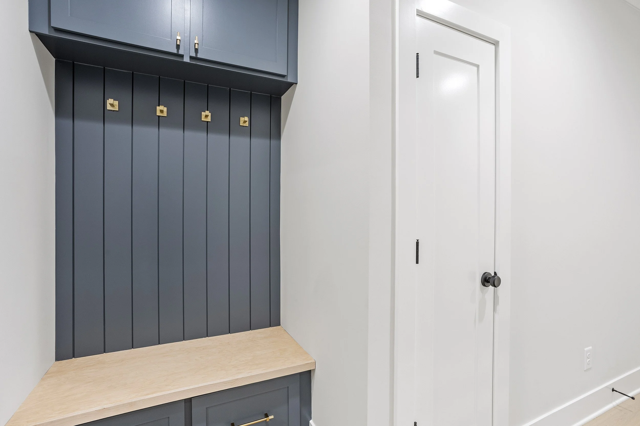 A modern interior space showing a section of a wall with a built-in dark blue cabinet with gold handles, a light wooden countertop, a white wall with trim, and a white door with a black doorknob.