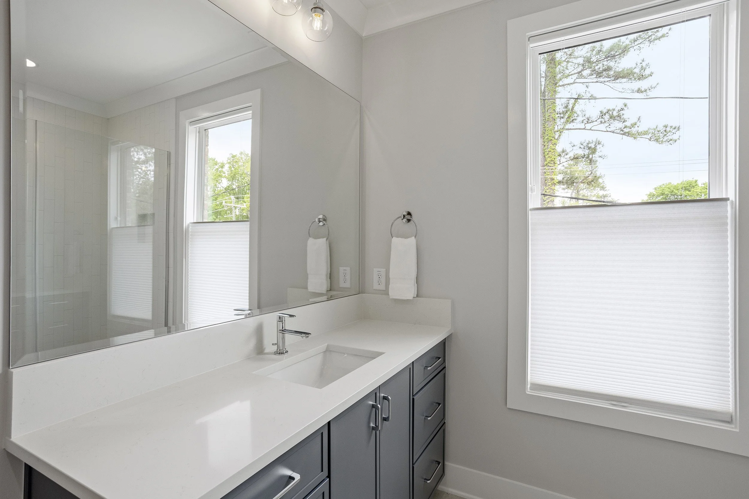 A modern bathroom with a white countertop, dark lower cabinets, large window with white blinds, mirror, and a small glass-enclosed shower in the corner. Outside the window, trees are visible.