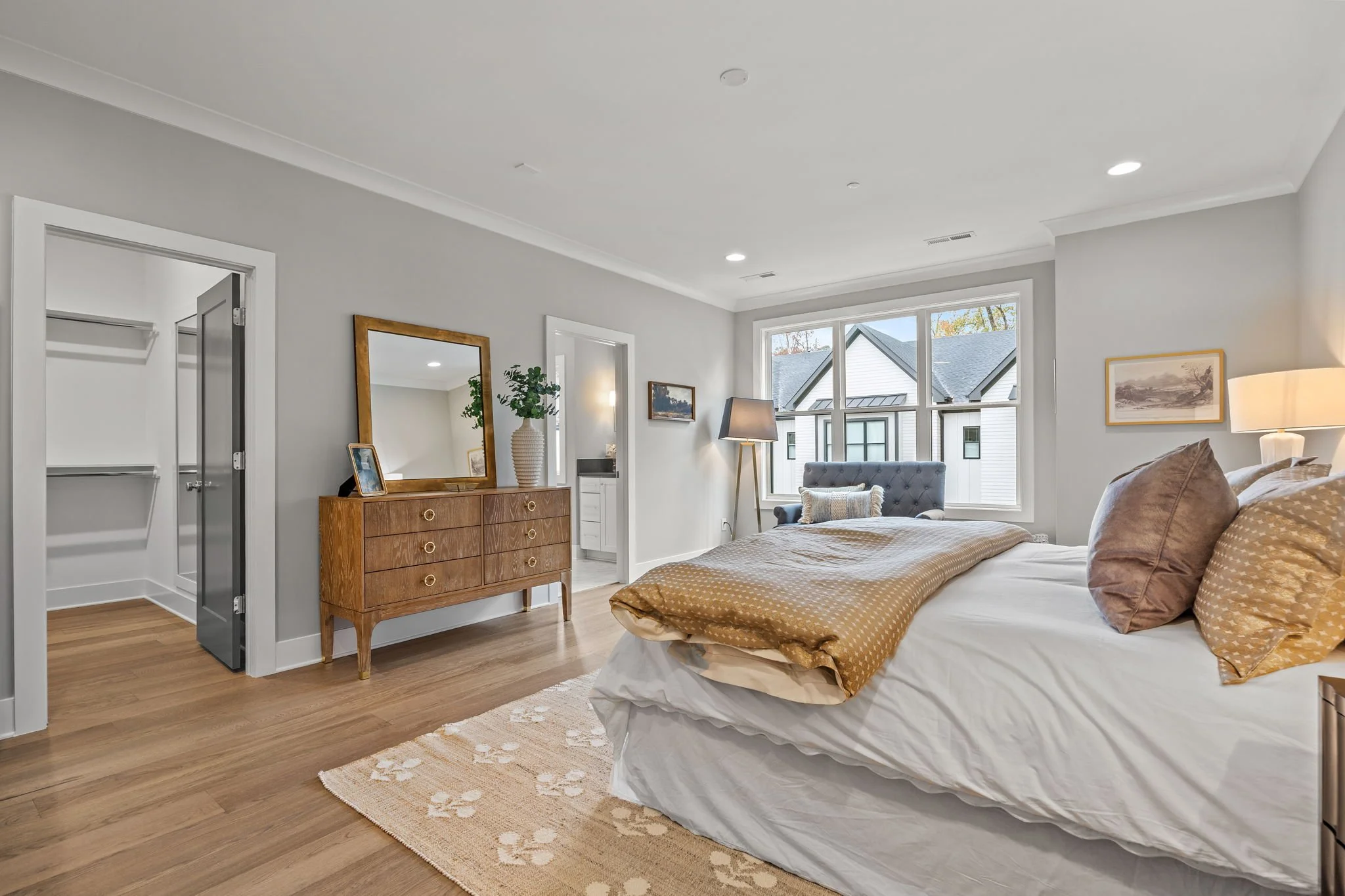 A modern bedroom with a bed, a wooden dresser, a large mirror, a potted plant, framed artwork, and a window showing neighboring houses.