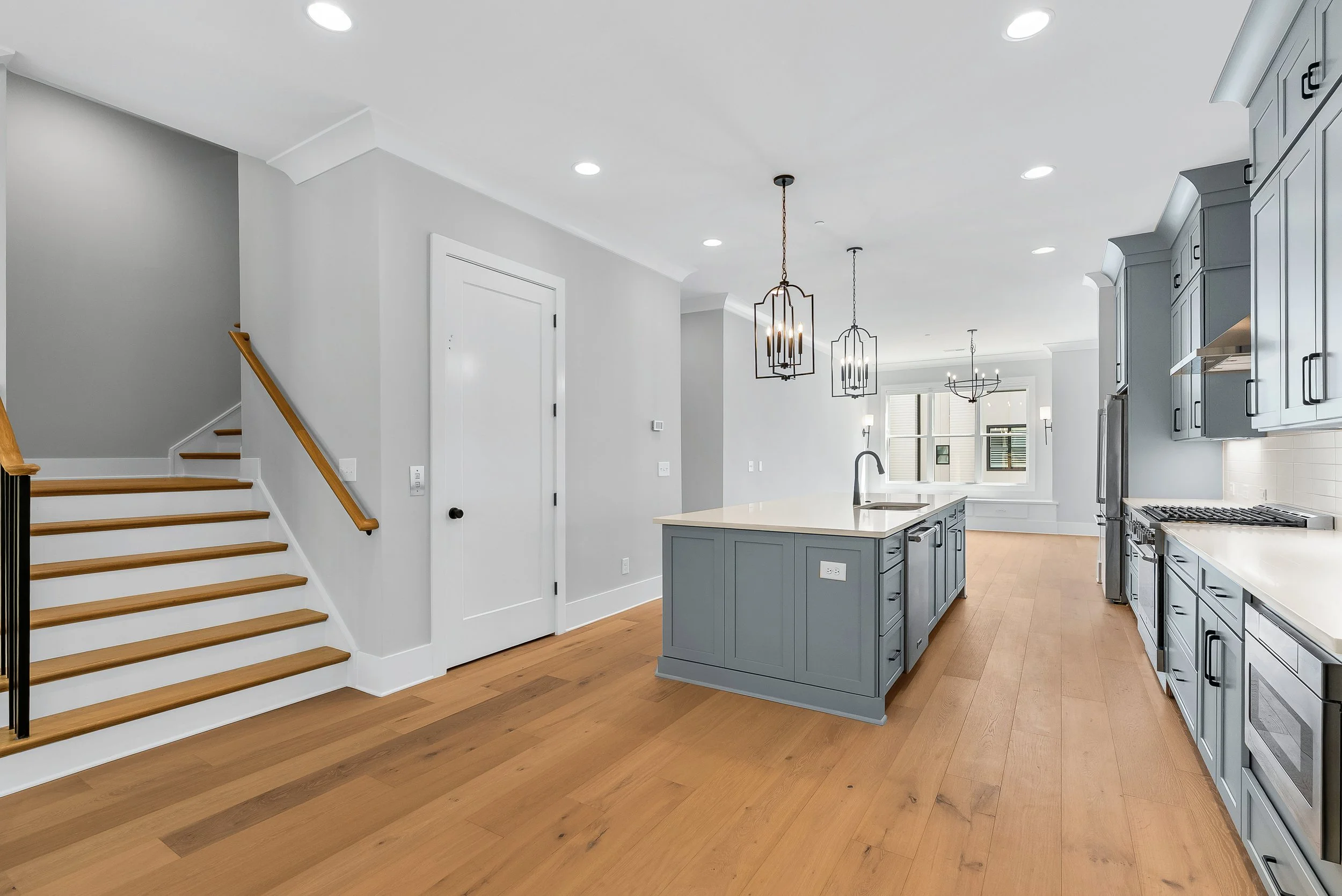 Open-concept kitchen with gray cabinetry, white countertops, a central island, hardwood flooring, modern black pendant lights, and a staircase with wooden handrail.