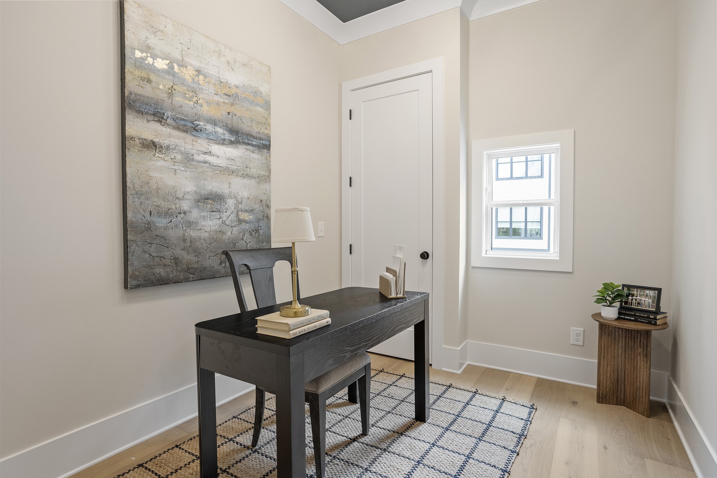 A cozy corner of a room with a small wooden table, decorative items, a framed photo, and a window with a grid pattern on the glass. There's a large abstract painting on the wall and a woven side table with a potted plant next to it. The room has ligh