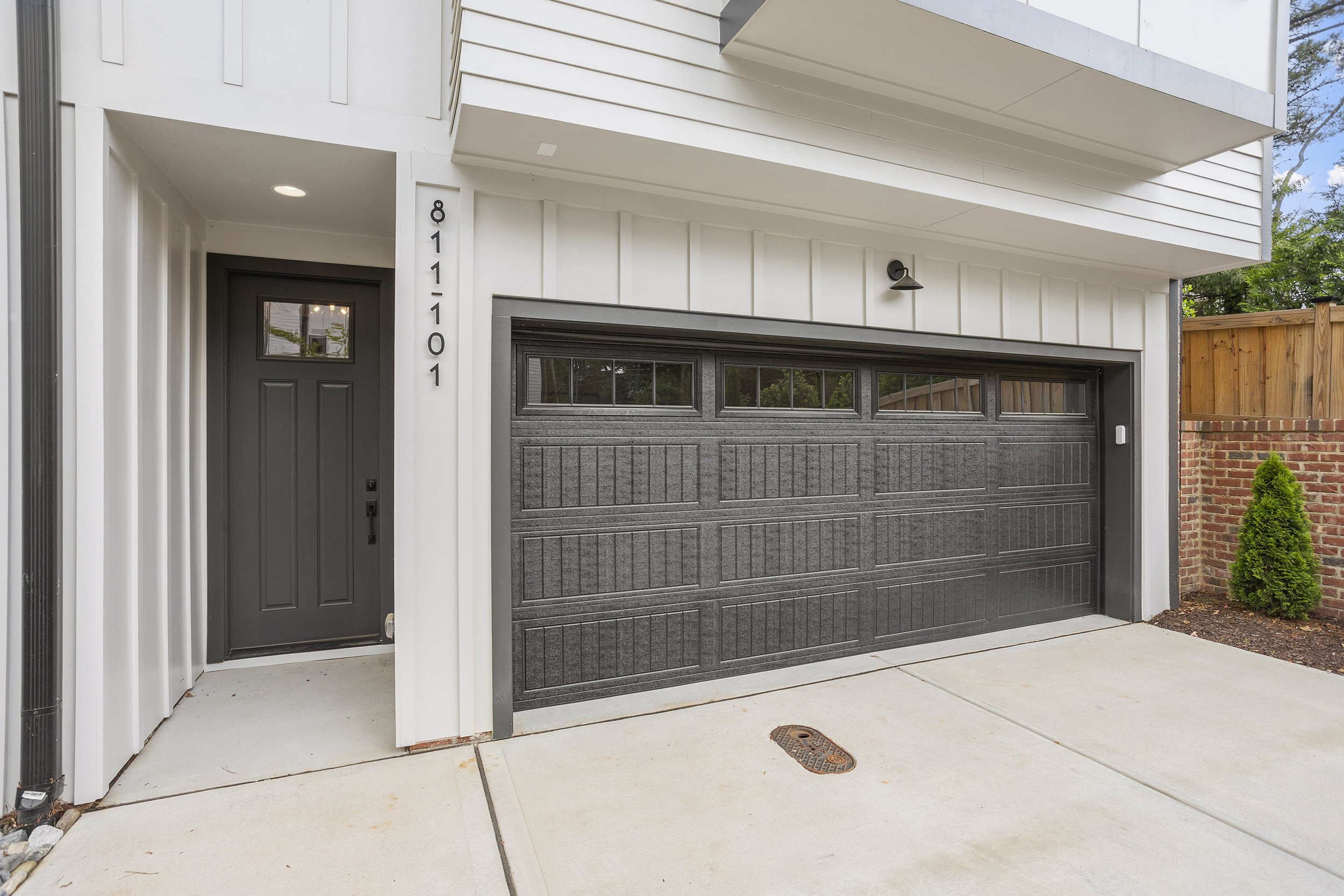 Front view of a modern house entrance with a black garage door, white exterior walls, and a dark gray front door, with a house number 811-10 on the wall, surrounded by a concrete sidewalk.