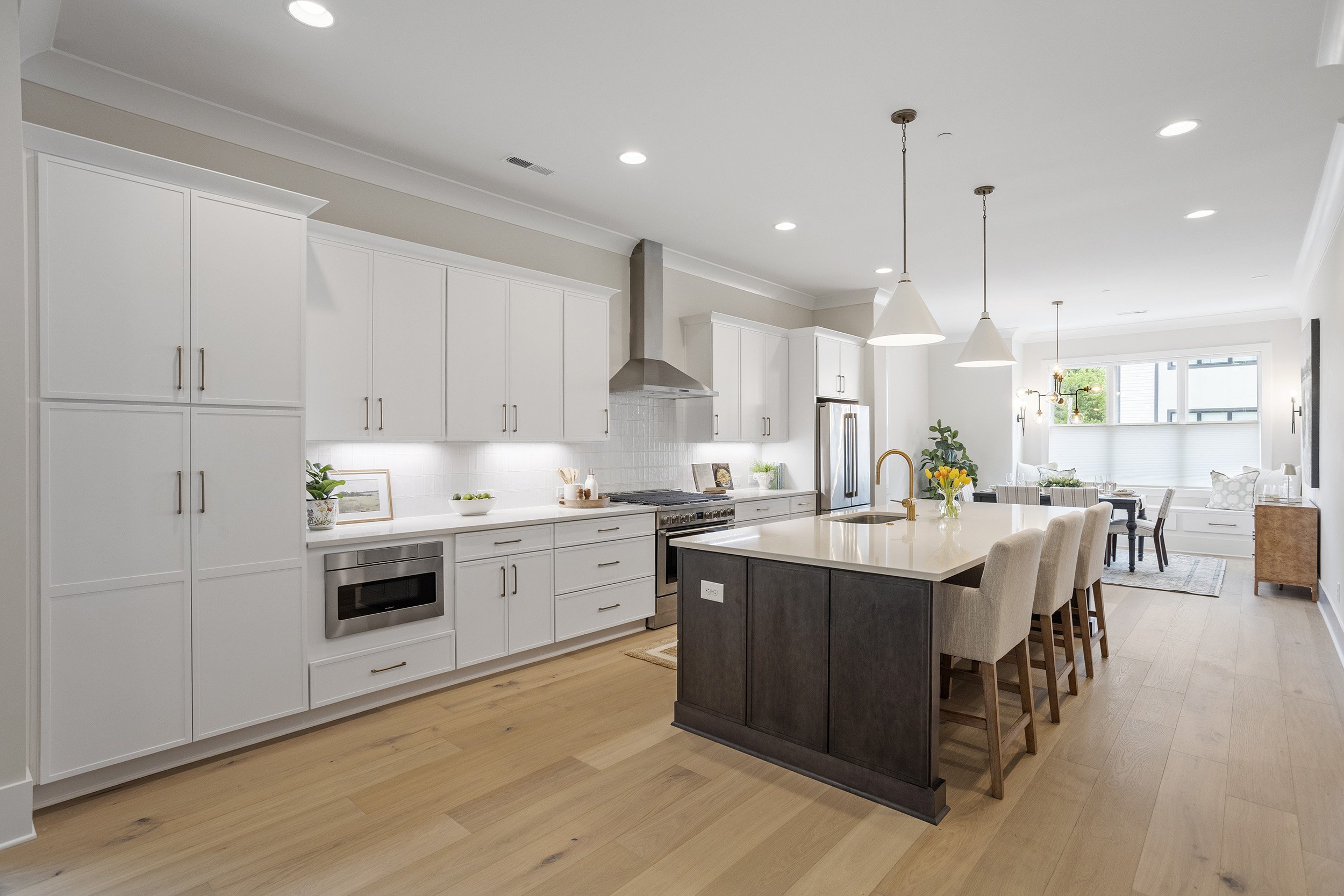 Modern open-concept kitchen and dining area with white cabinets, stainless steel appliances, a large island with seating, pendant lighting, and a bright living space with large windows.