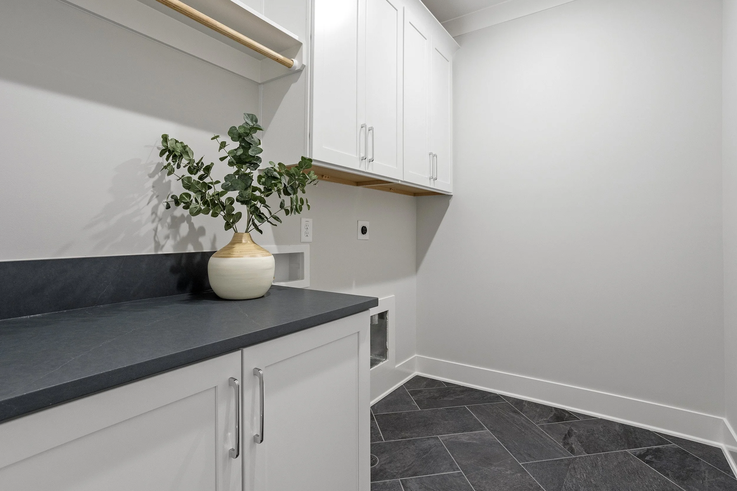 Interior view of a kitchen with white cabinets, black countertop, and dark tiled floor. There is a green plant in a beige and cream vase on the countertop. A white wall with electrical outlets and an open space under a cabinet is visible.