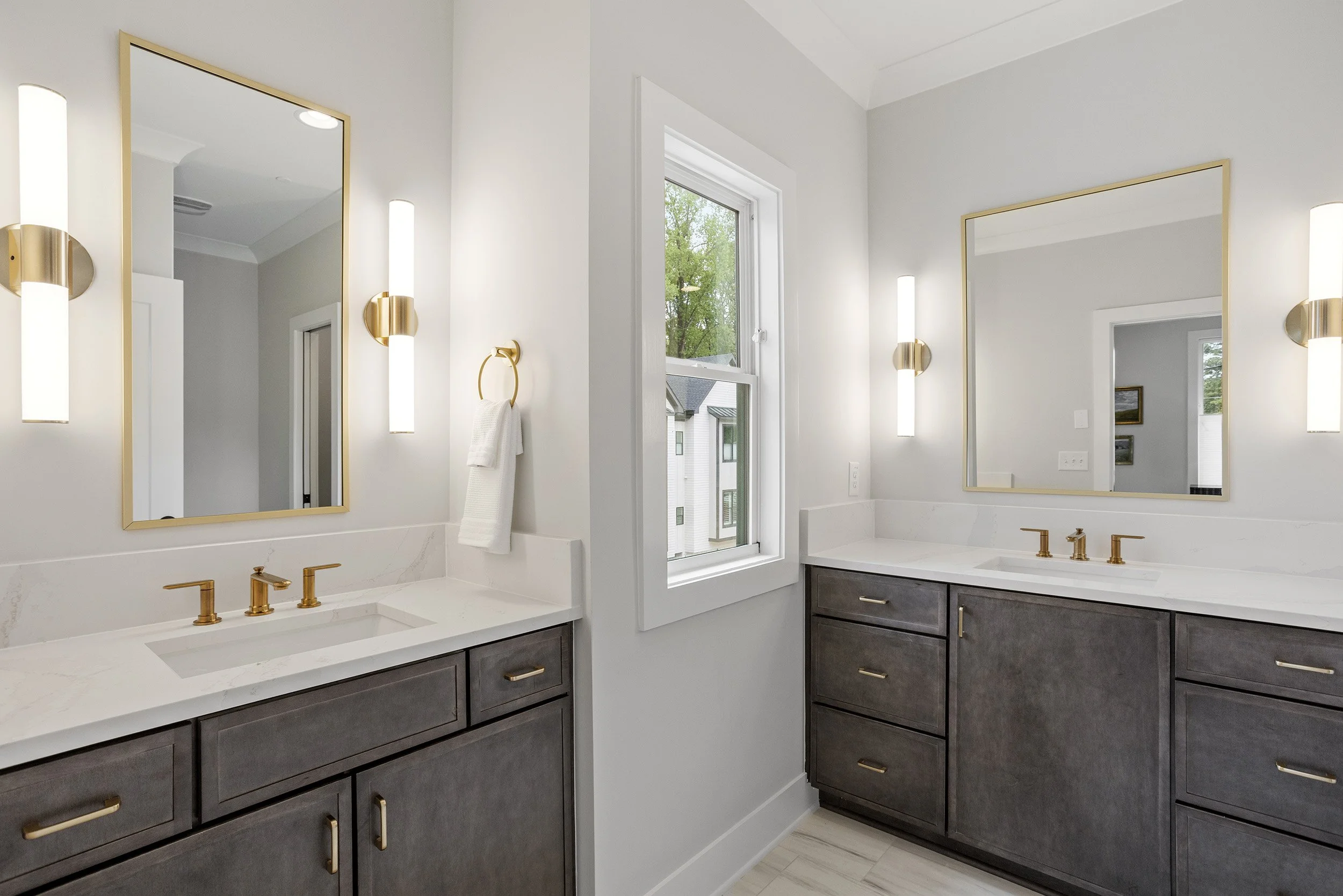 Modern bathroom with dark wood cabinets, white countertops, gold fixtures, large mirrors, white walls, and a window showing greenery outside.