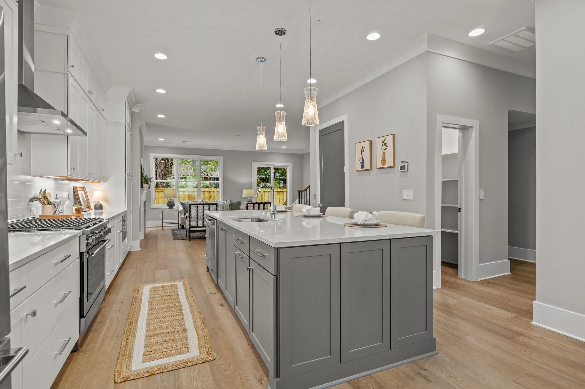 A modern, bright kitchen with white cabinets and a large central island with a gray base and white countertop. Pendant lights hang above the island. There are wooden floors, and a rug runs along the floor leading to a living area with large windows a