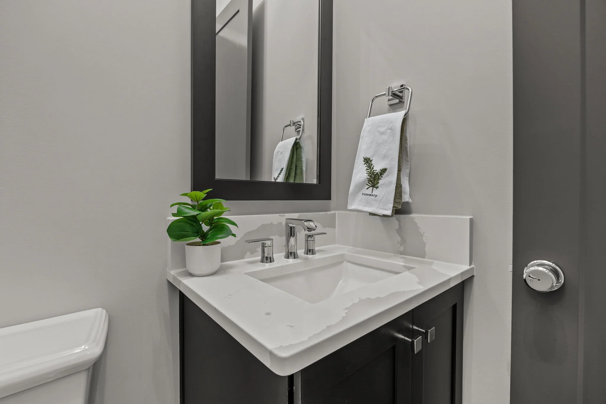 A bathroom sink vanity with a white marble countertop, a rectangular mirror, a silver faucet, a potted green plant, a towel with a botanical print labeled 'rosemary', and a partial view of a toilet.