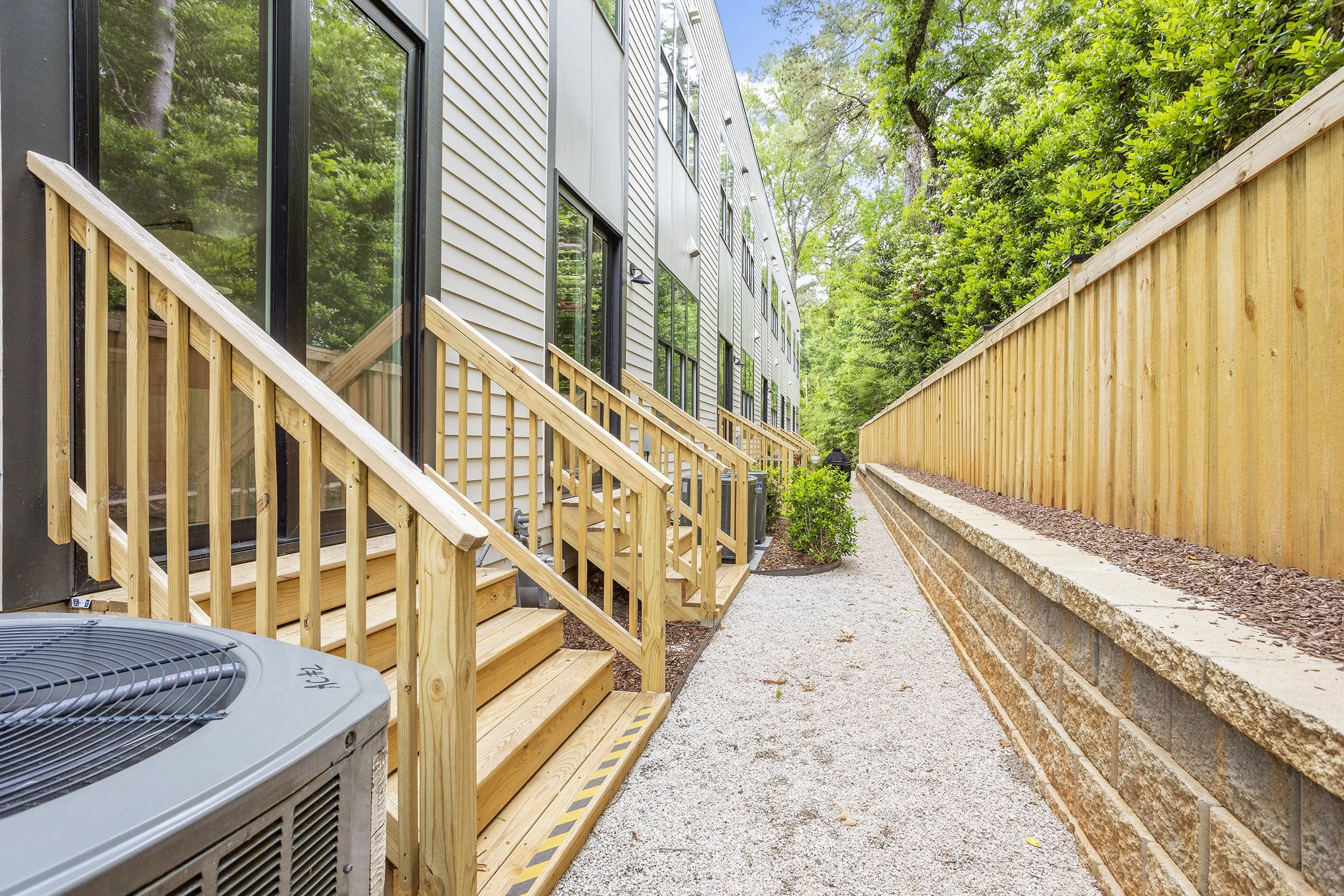 A side view of a modern multi-story apartment building with large glass windows, a small backyard with a wooden staircase, a gravel pathway, and a tall wooden fence with lush green trees.
