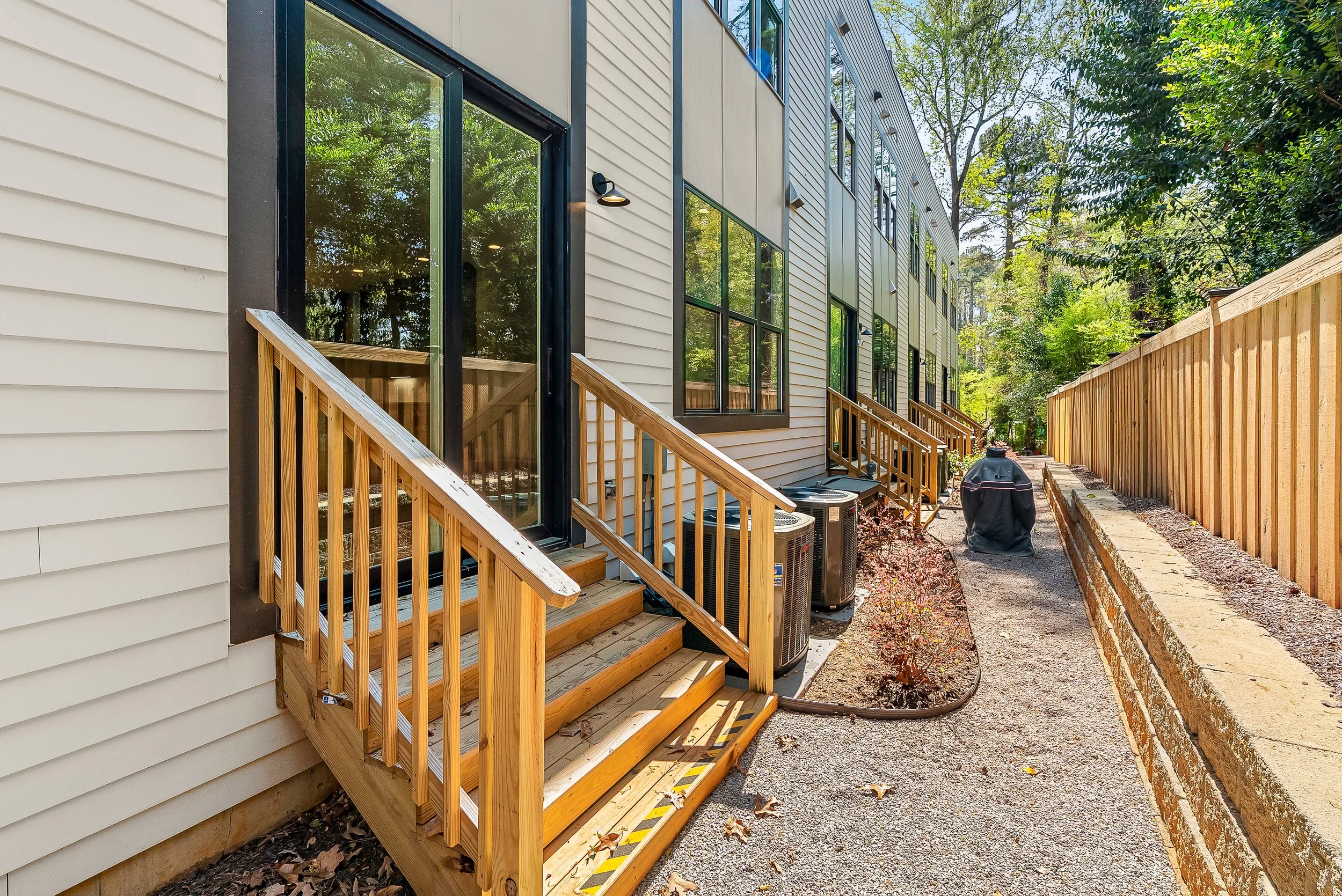 Newly built wooden stairs leading up to the entry of a modern apartment building with siding and large windows, surrounded by trees and a wooden fence.