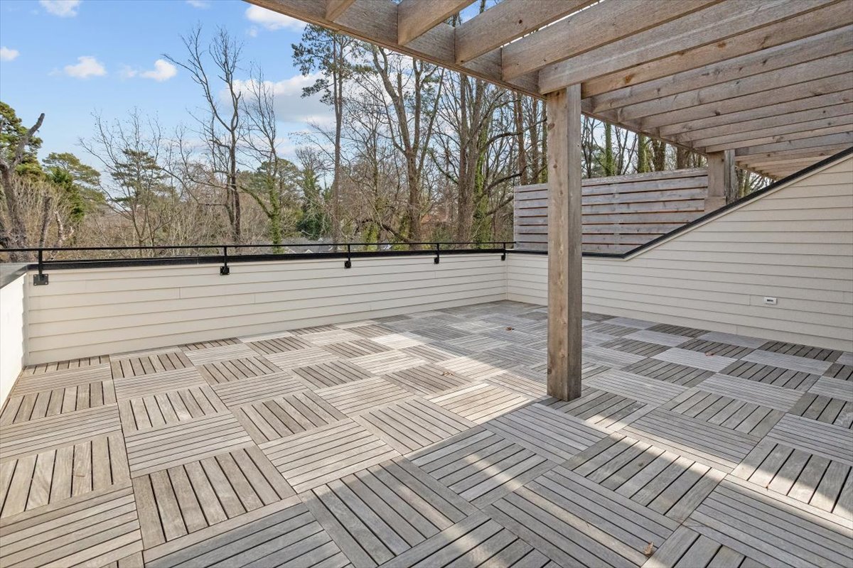 Outdoor wooden deck with a partially covered roof, surrounded by a black railing and a white wall, overlooking a wooded area with leafless trees.