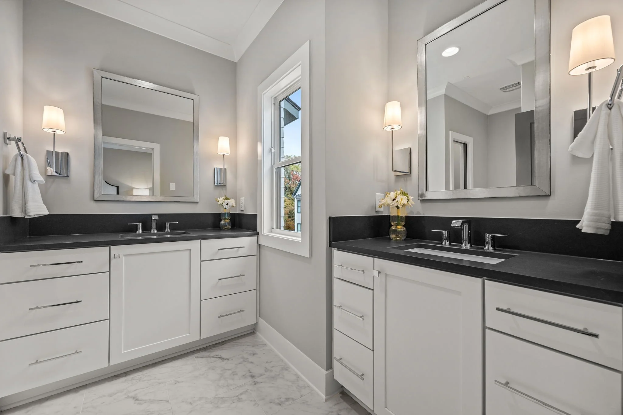 Modern bathroom with gray walls, white cabinetry, black countertops, large mirrors, wall-mounted lamps, a window, and a vase with white flowers.