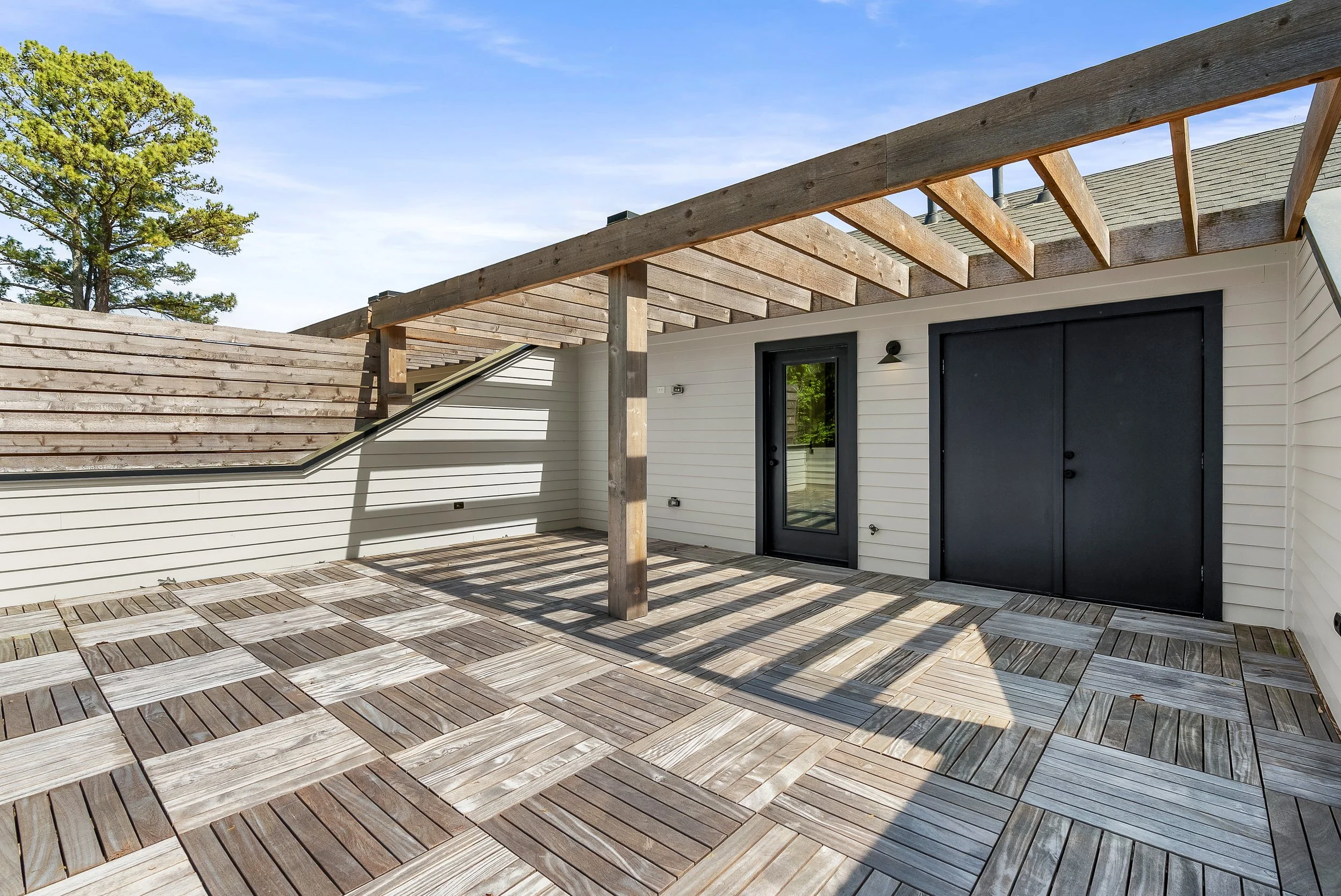 A spacious balcony with a wooden lattice overhead, a sliding glass door with black trim, cream-colored siding, and a floor made of interlocking wood tiles, with a large tree and blue sky in the background.