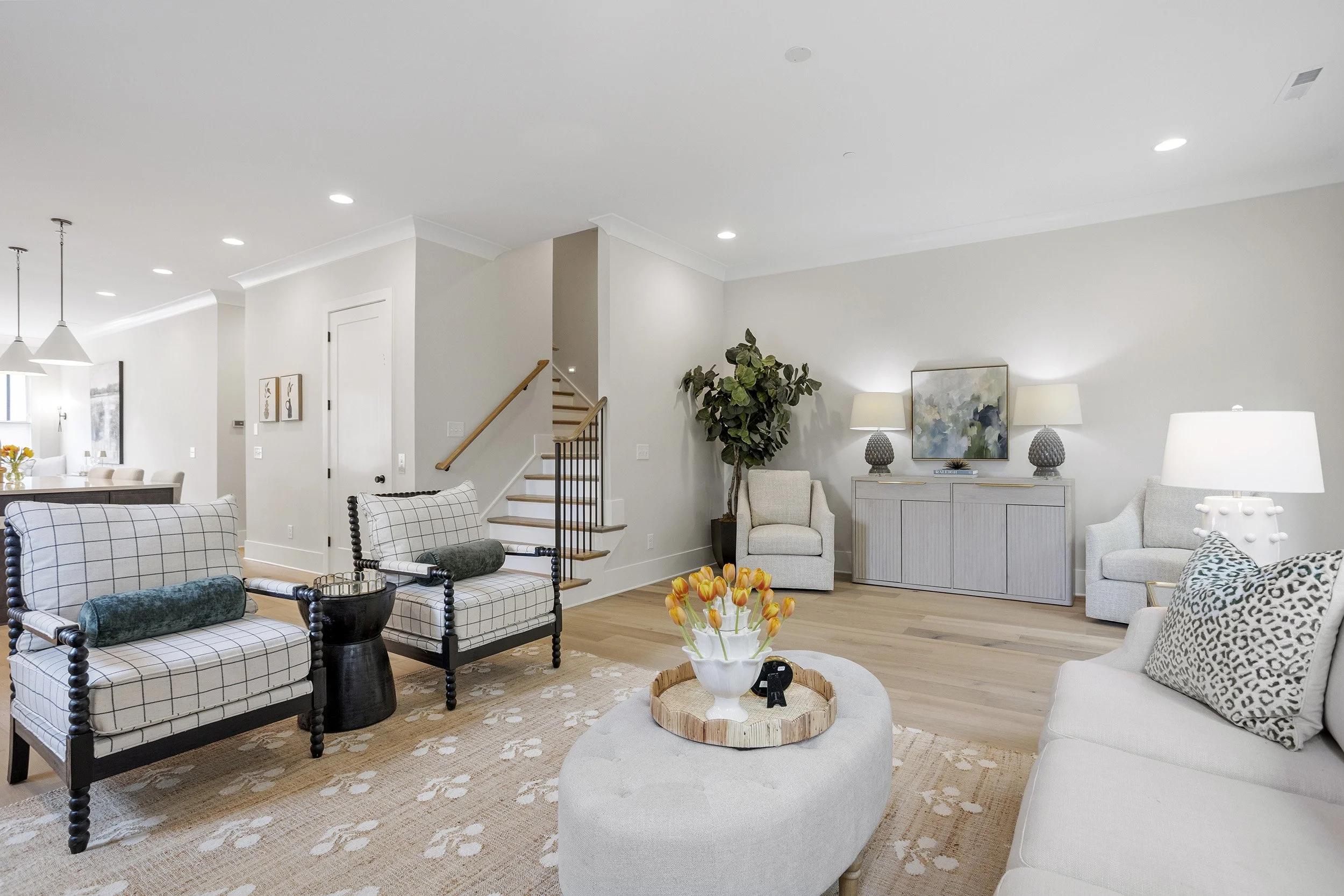 Living room with white walls and wooden flooring, featuring patterned and plain chairs, a coffee table with a vase of orange flowers, a potted plant, and wall art.