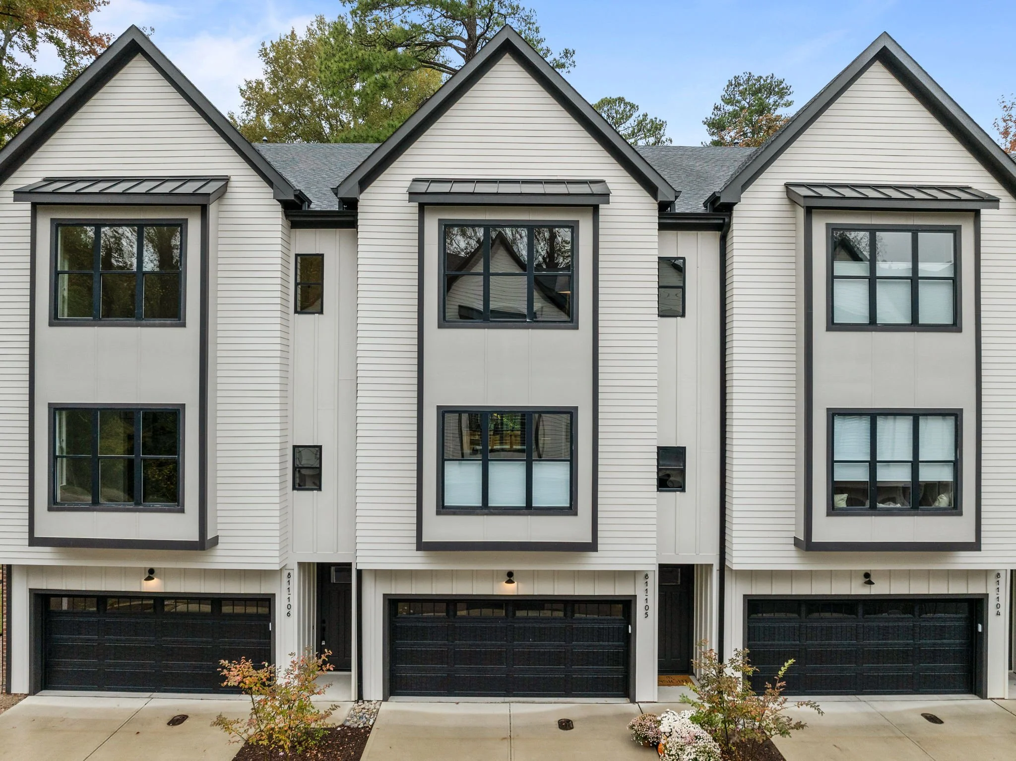 A modern multi-story residential building with white siding and black-framed windows, three front-facing gables with black awnings, and black garage doors at street level. Small landscaped front area with plants and flowers.