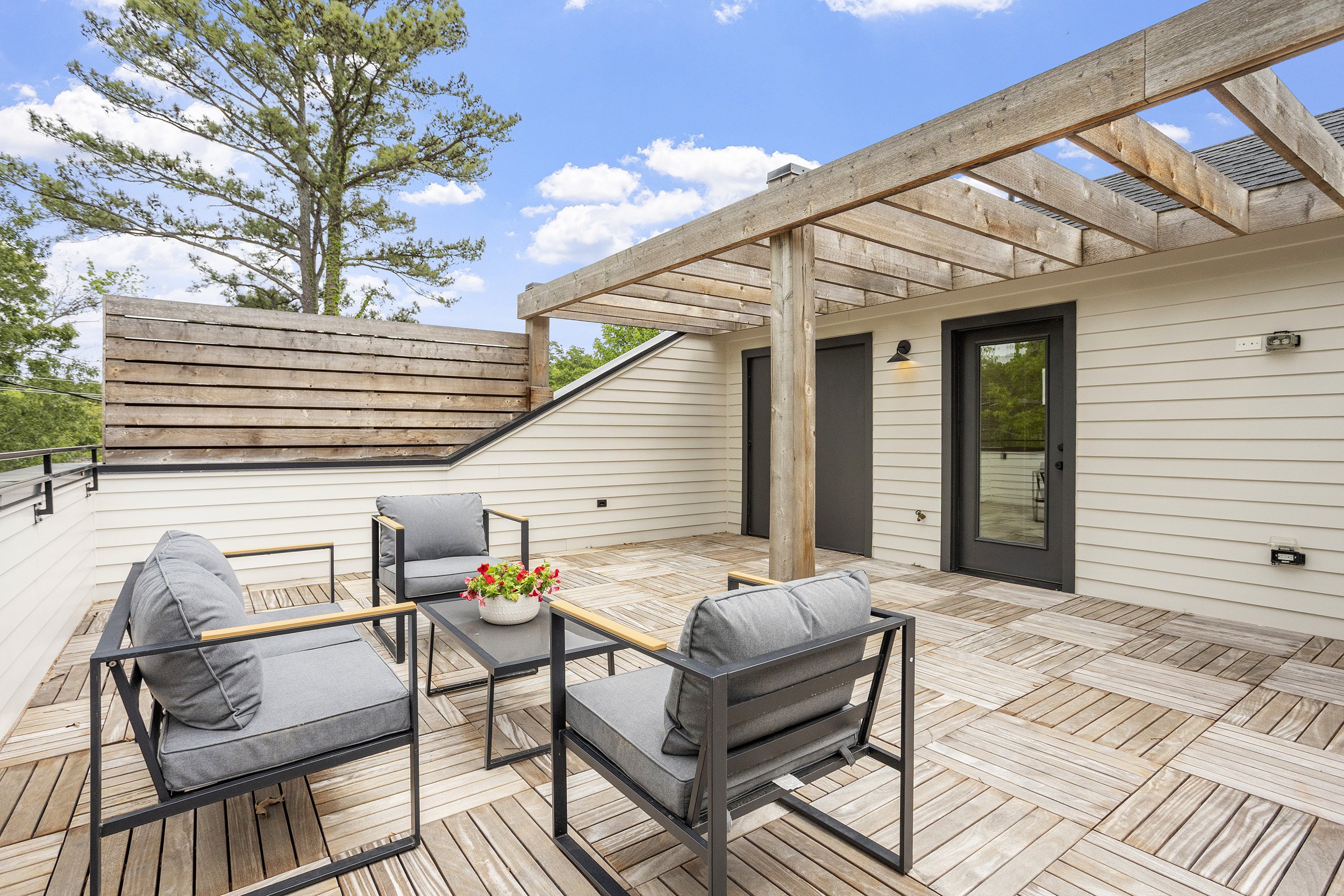 A rooftop patio with outdoor furniture, including four chairs with dark frames and gray cushions, a black table with a potted red flower, wooden flooring, a white building with black doors, a wall-mounted light, and a partially constructed wooden per