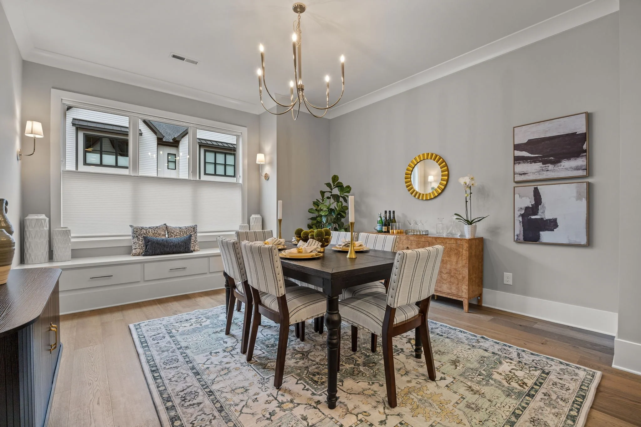 A dining room with a dark wooden table, six striped upholstered chairs, a gold candle holder, and a decorative tray with drinks. There is a built-in window seat with pillows under a large window, and wall art with abstract paintings. The room has a c