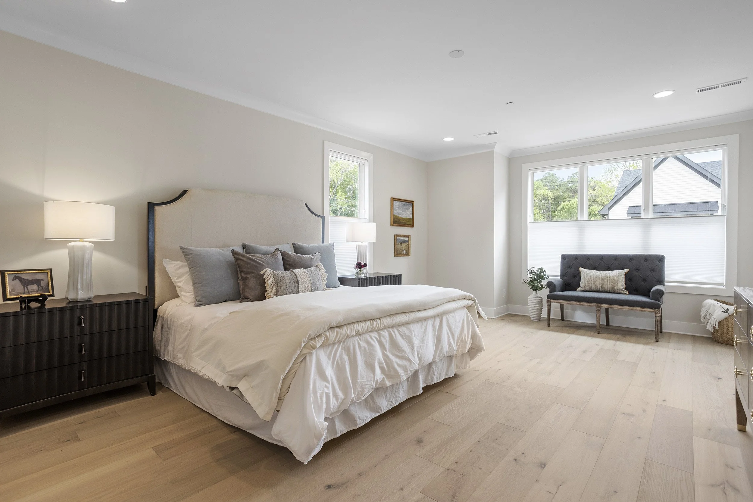 A bedroom with a bed, nightstand, lamps, and seating area by large windows with a view of trees and a neighboring house.