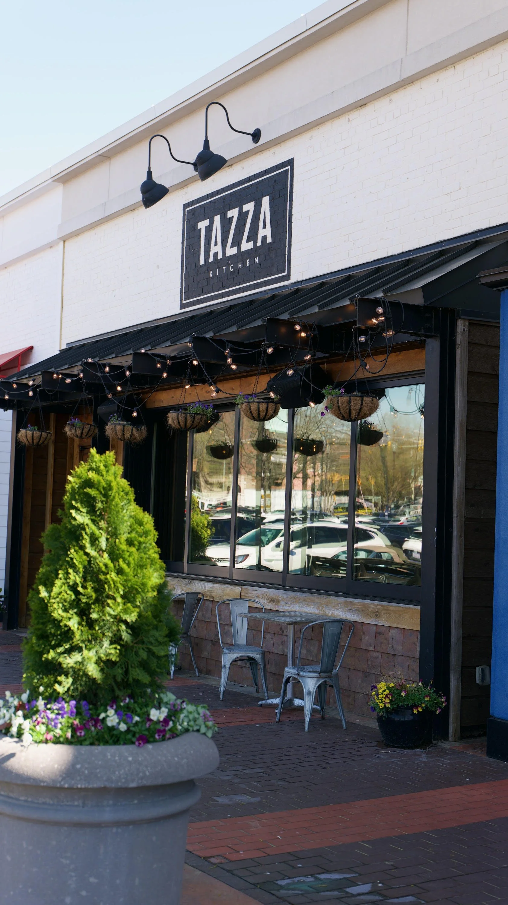 Exterior view of Tazza Kitchen with a white brick wall, black window frames, and outdoor seating area featuring chairs, plants, and hanging string lights.
