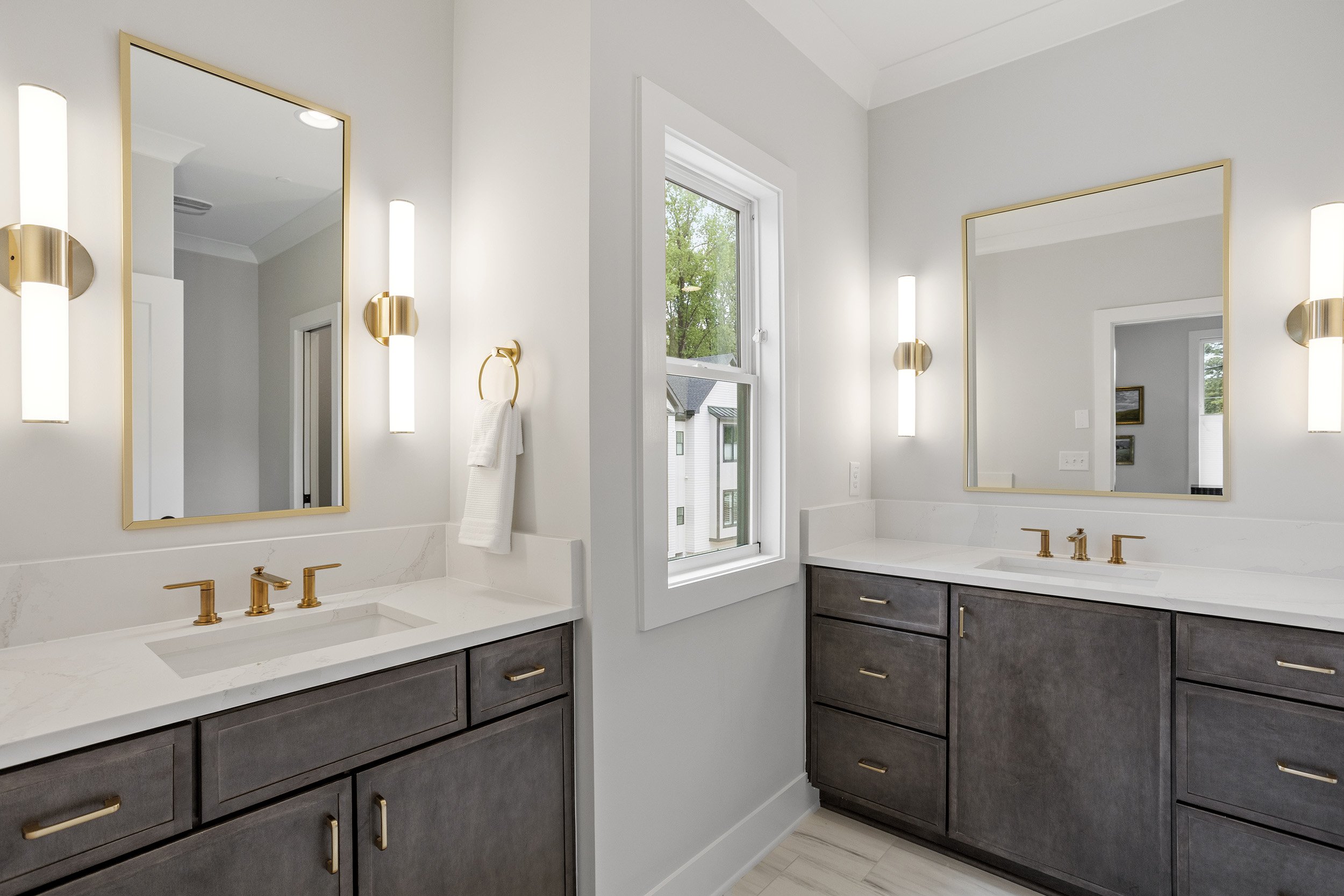 A modern bathroom with double vanity sinks, gold fixtures, large framed mirrors, and a window showing trees outside.