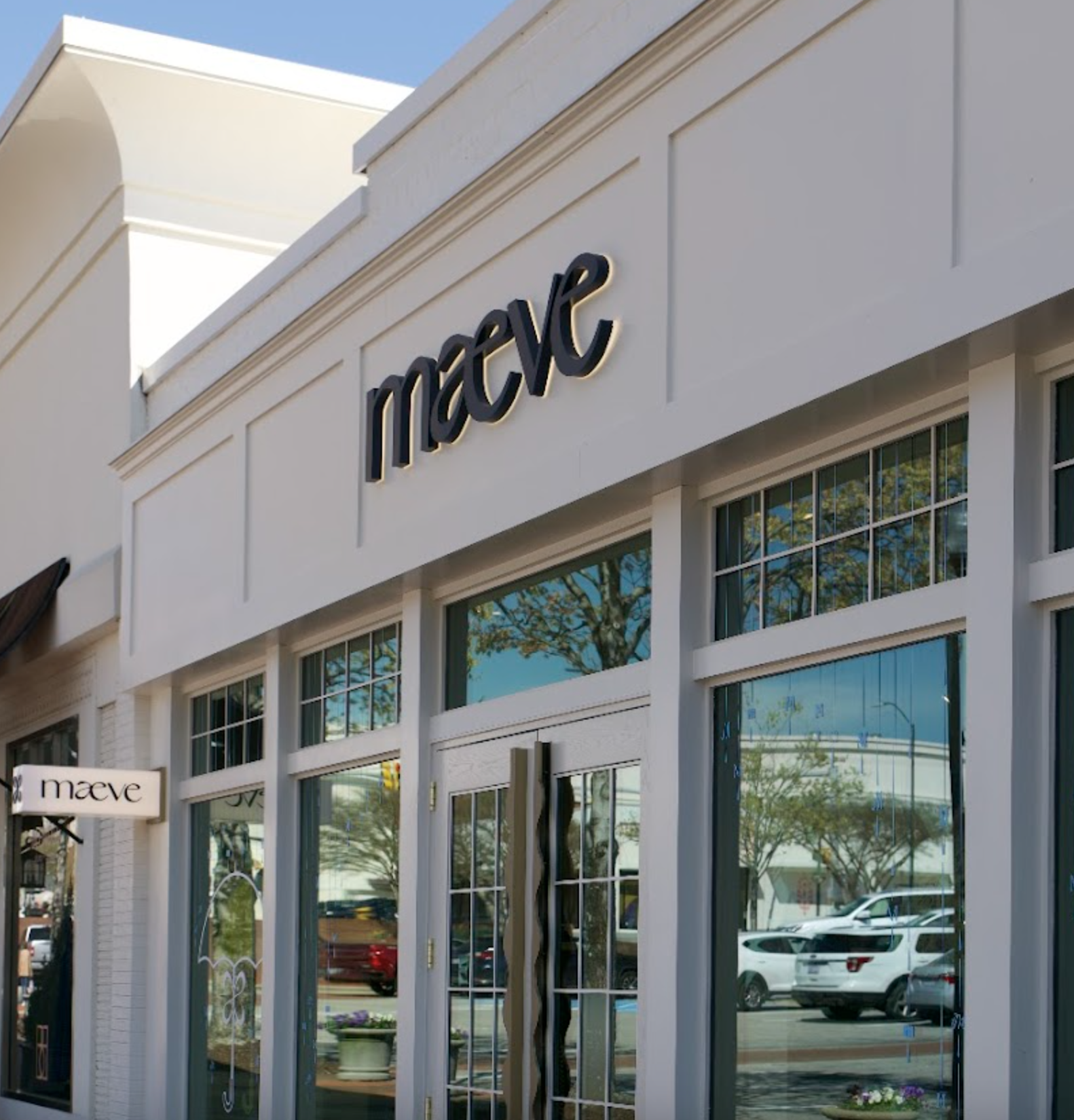 The storefront of a retail store named 'Market' with large glass windows and white framing. There are reflections of trees and cars in the windows, and some smaller signs including one for 'maeve' are visible.