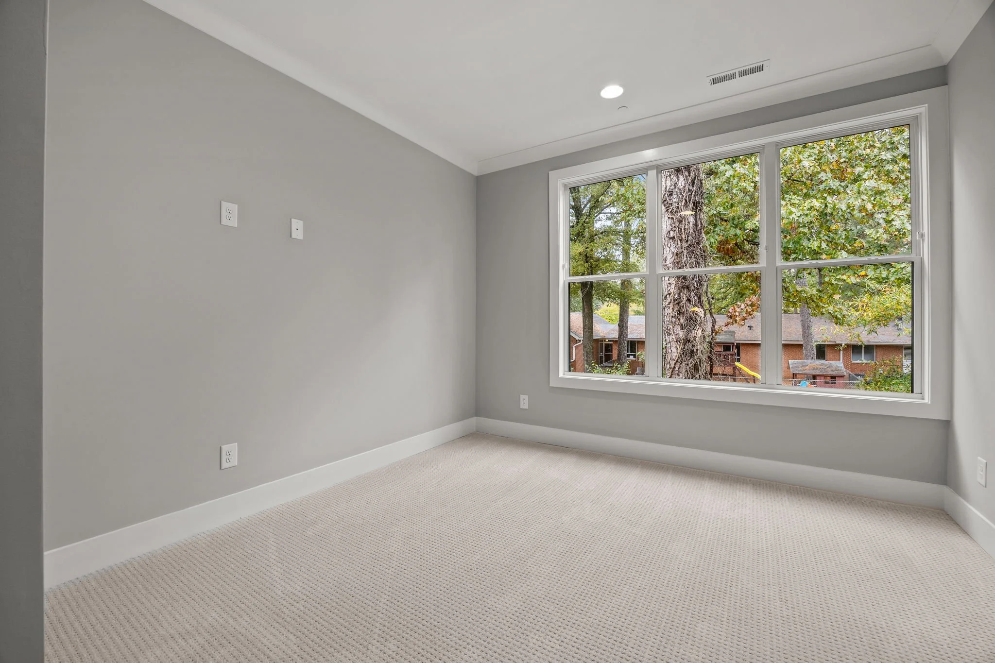 Empty room with beige carpeting, light gray walls, a large window showing trees outside, and ceiling lights.