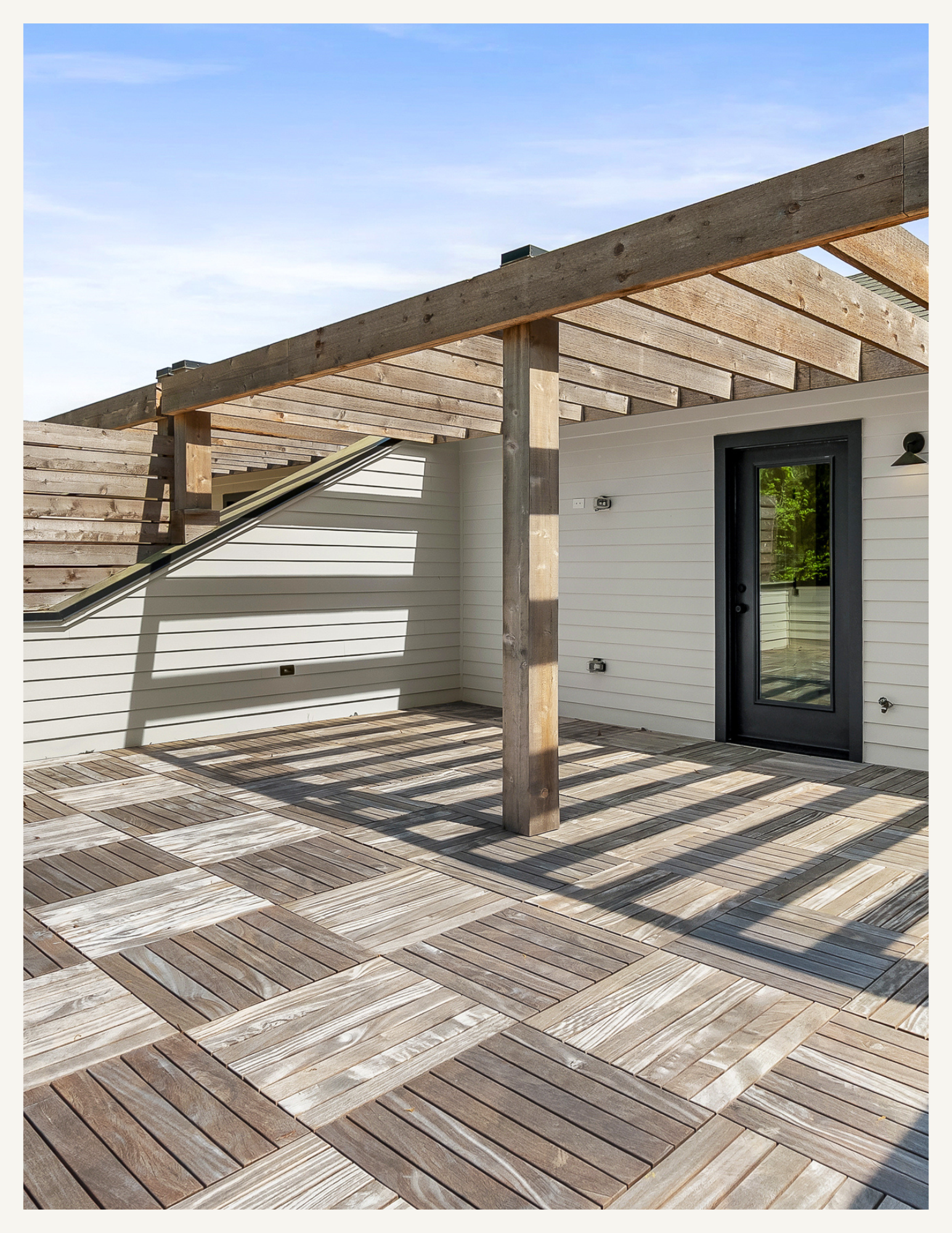 A patio with wood planks arranged in a diagonal pattern, part of a modern outdoor space, with a house wall, door, and wooden pergola structure casting shadows.
