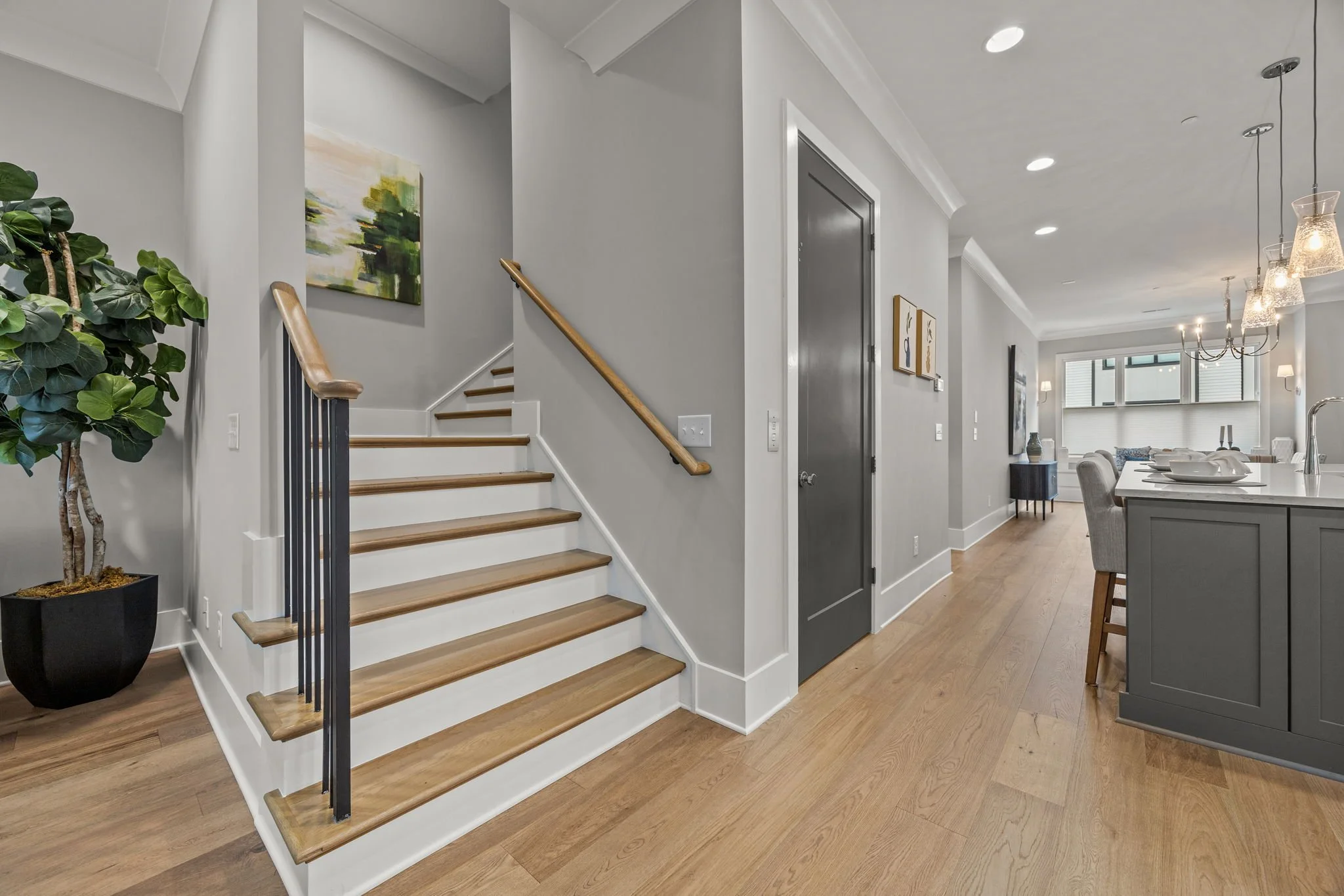 A modern open-concept living space with a staircase, a potted plant, framed artwork on the wall, and a dining area with a chandelier. The room features light-colored walls, hardwood floors, and large windows.
