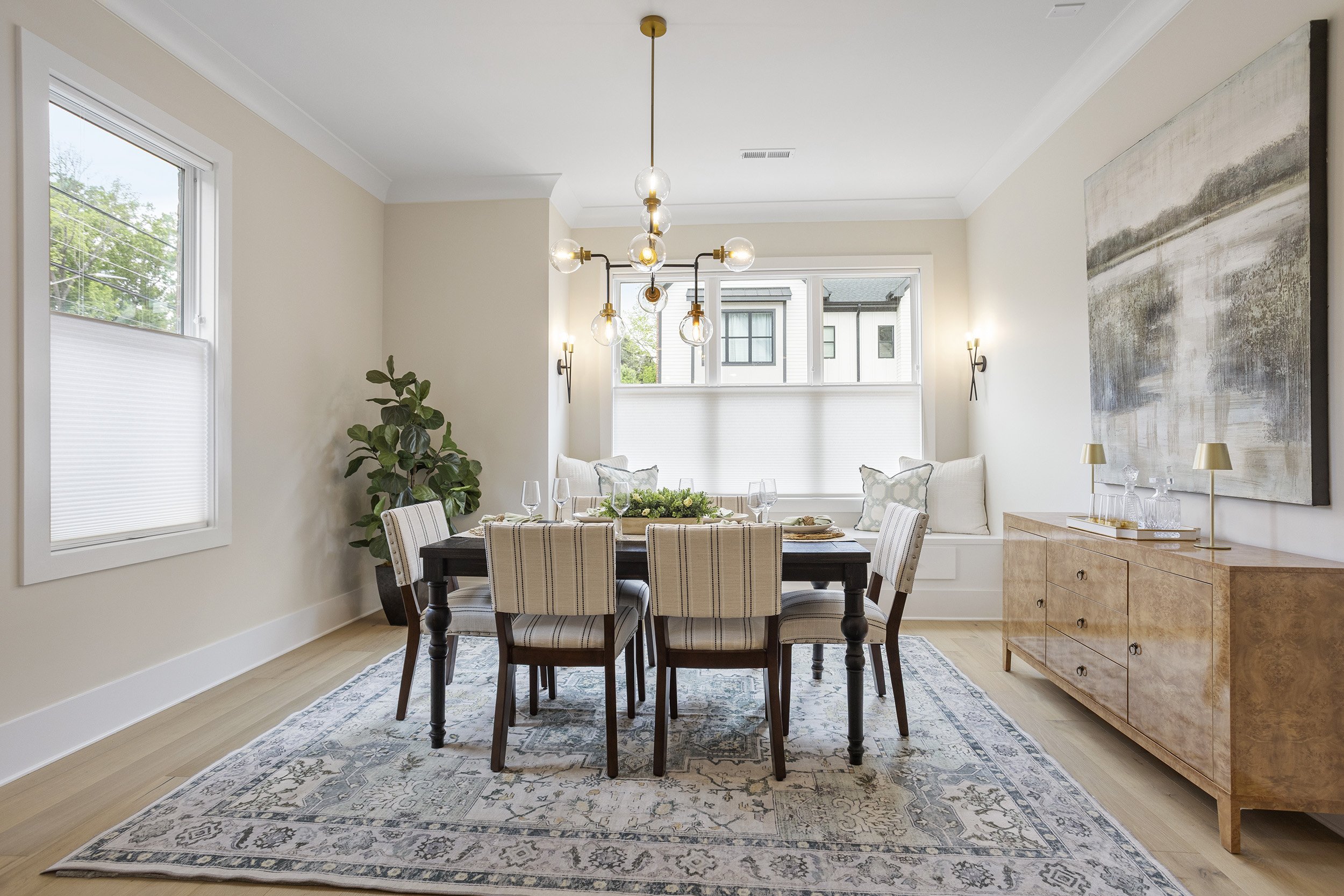 A dining room with a wooden table, white and striped upholstered chairs, a decorative table setting, a large window seat with cushions, a chandelier, outside views of neighboring houses, a patterned rug, a tall plant, a wooden sideboard with glassware and lamps, and abstract wall art.