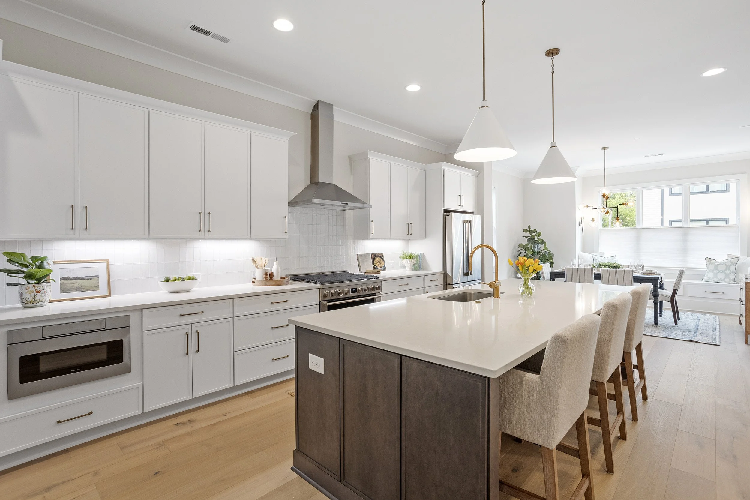 Modern kitchen with white cabinets, gray island, stainless steel appliances, pendant lighting, and dining area with large window and plants.