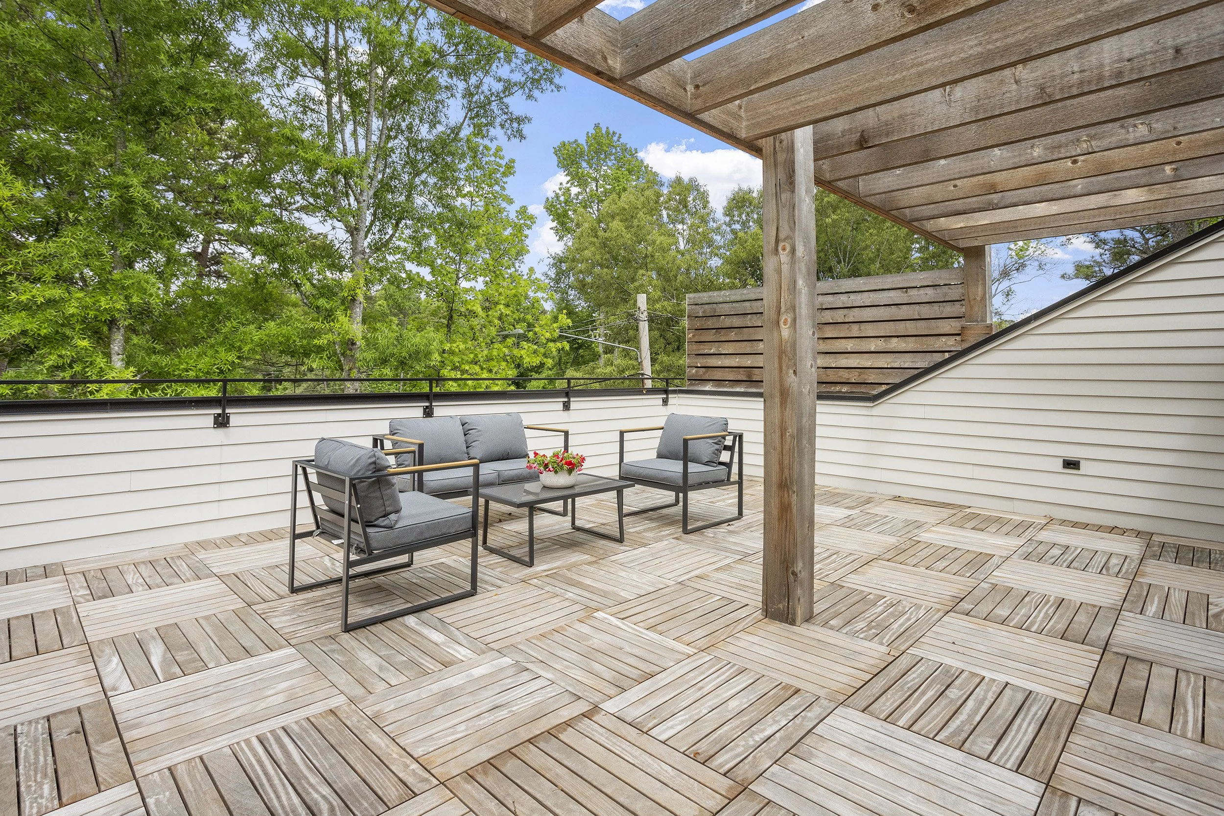 An outdoor patio with wooden flooring, a seating area with a sofa and two chairs, a coffee table with a flowerpot, surrounded by green trees and blue sky, partially shaded by a wooden pergola.
