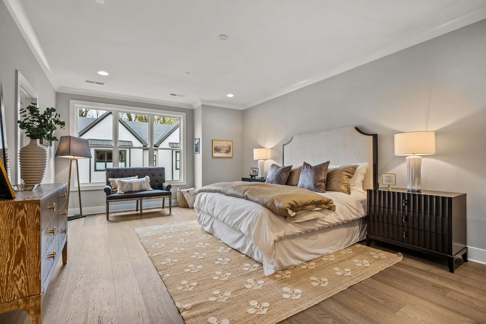 A cozy bedroom with a large bed featuring beige and brown patterned bedding, wooden nightstands with lamps, a window with a view of neighboring houses, and decorative plants and artwork in a modern, neutral-colored interior.