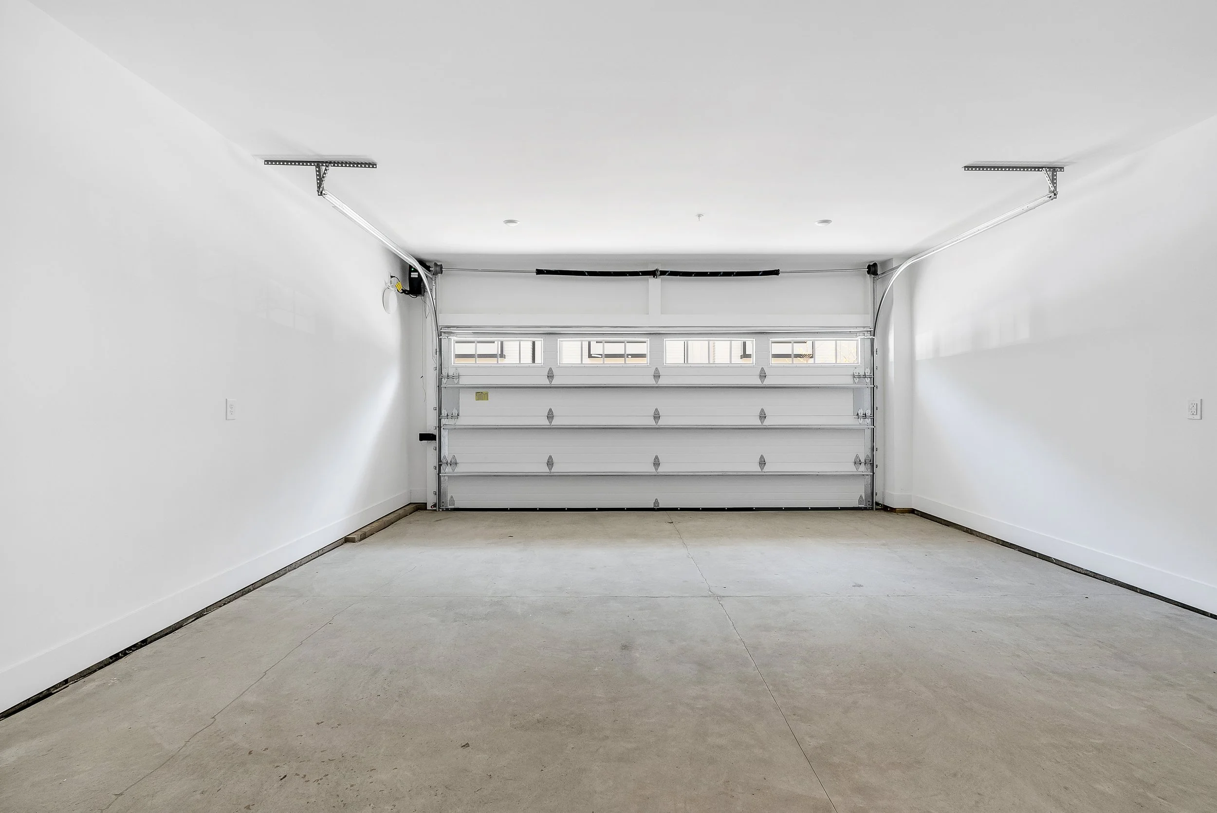Empty garage with white walls, concrete floor, and a closed white garage door. Wiring and garage door opener are visible on the ceiling.