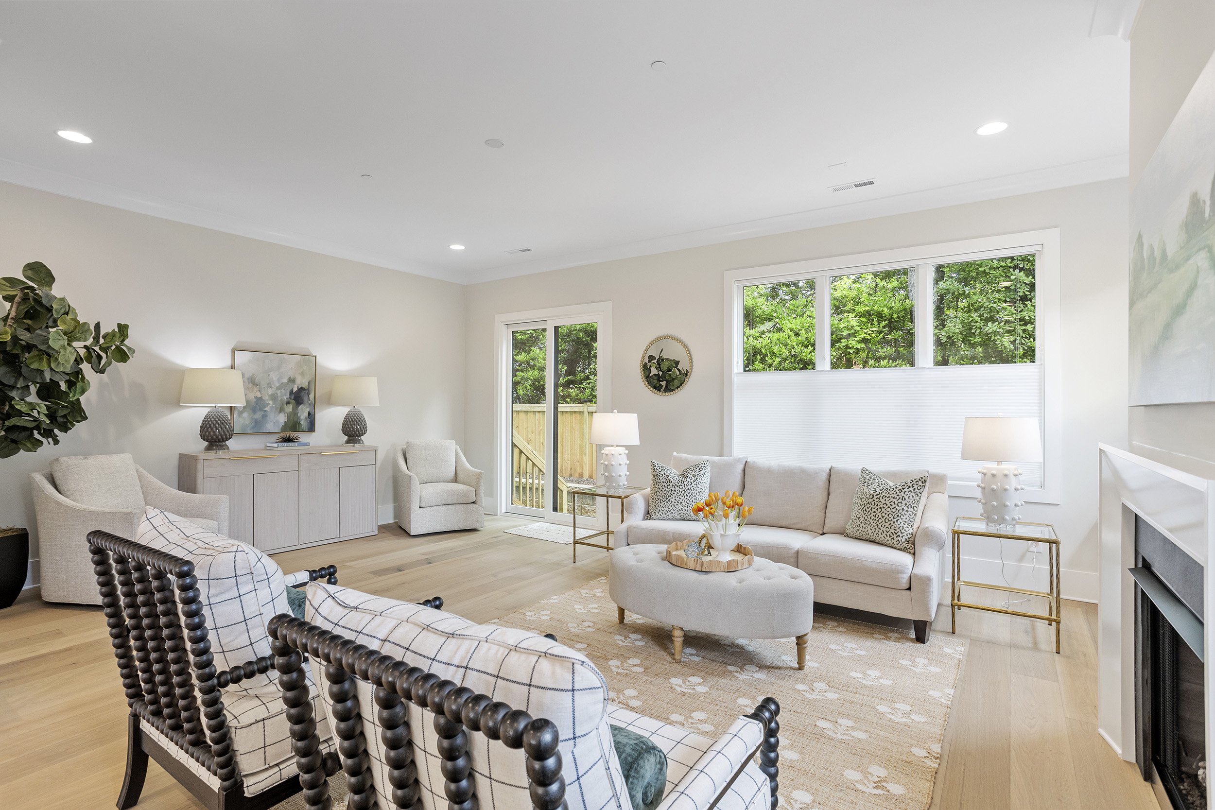 A spacious living room with beige walls, a large window showing greenery outside, white ceiling with recessed lighting, a grey sideboard with lamps, a armchair, a beige sofa with patterned pillows, a ottoman with a tray of flowers, and a patterned ru