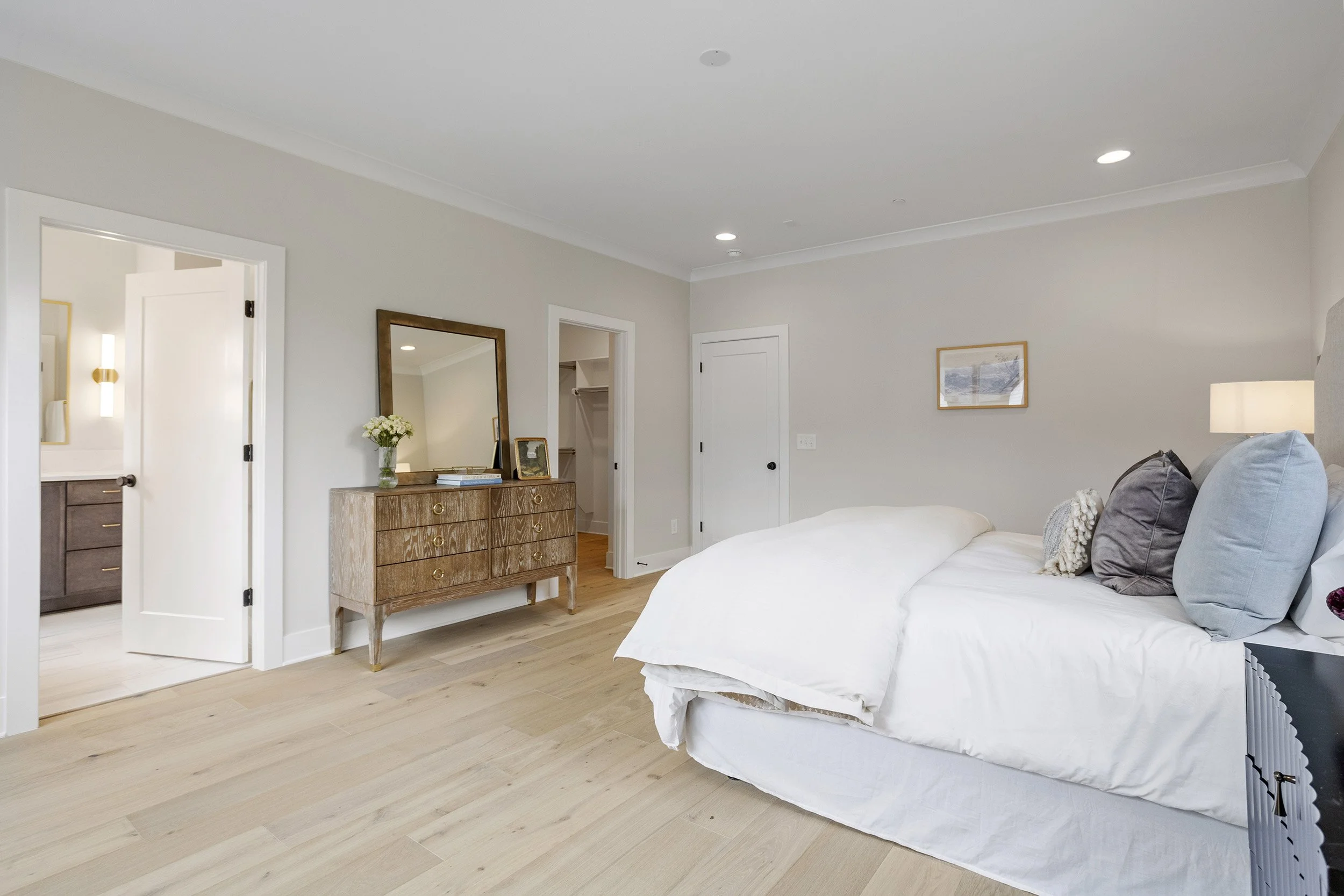 A bedroom with a white bed, pillows, a wooden dresser with a mirror and flowers, and a framed picture on the wall. There are closed doors and light wood flooring.