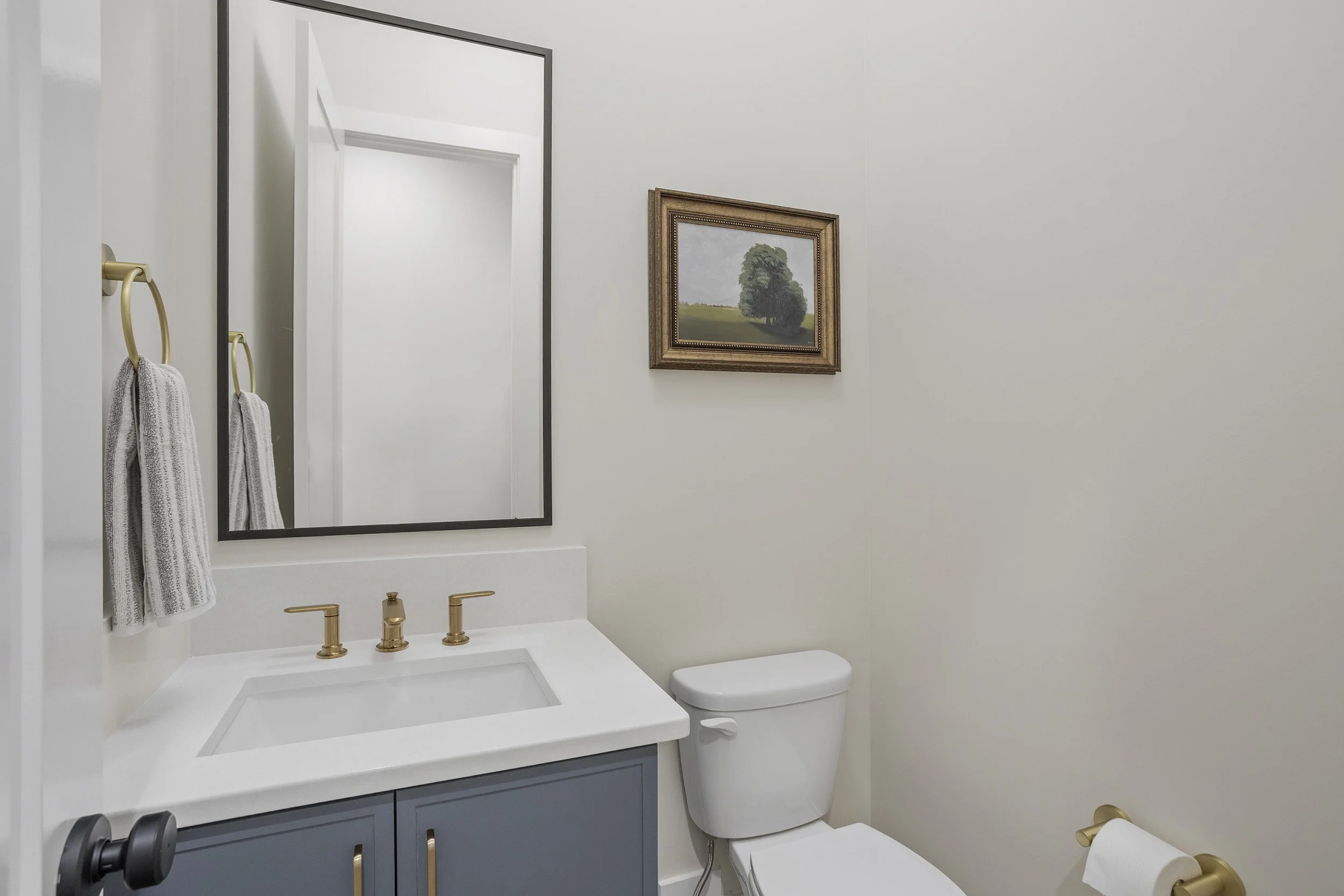 A small bathroom with a white vanity with a square sink, gold faucet, and blue cabinet. A large rectangular mirror hangs above the sink. Gold towel hooks on the wall hold two white towels. A framed landscape painting of trees and a green field is on 