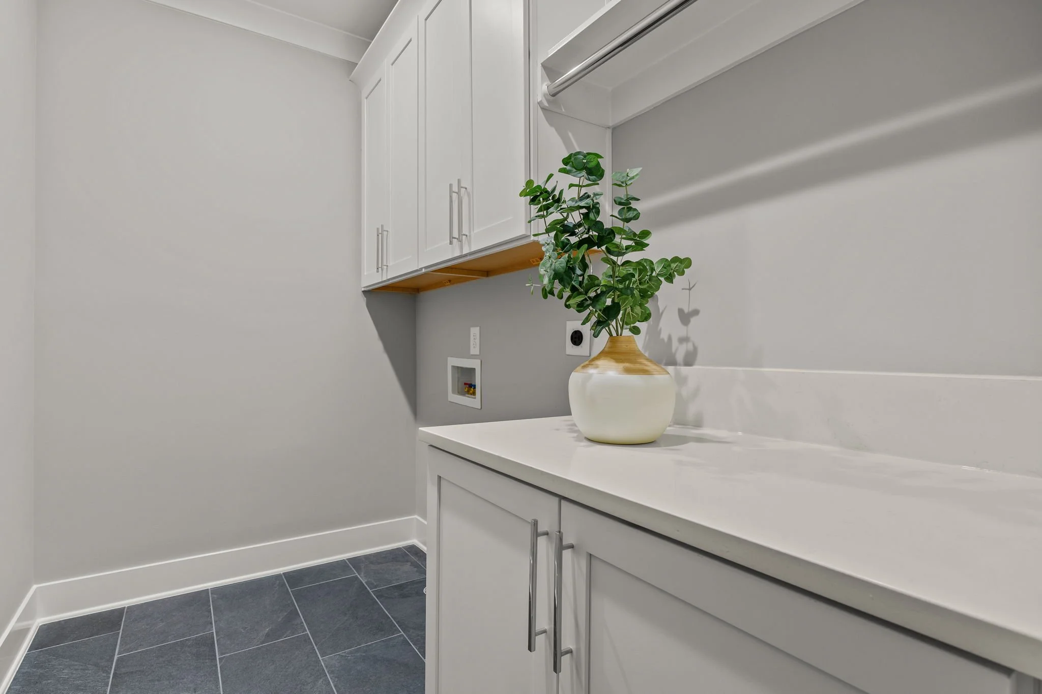 A white countertop with a vase of green leafy plant, white cabinets above, gray tiled floor, and plain white walls.